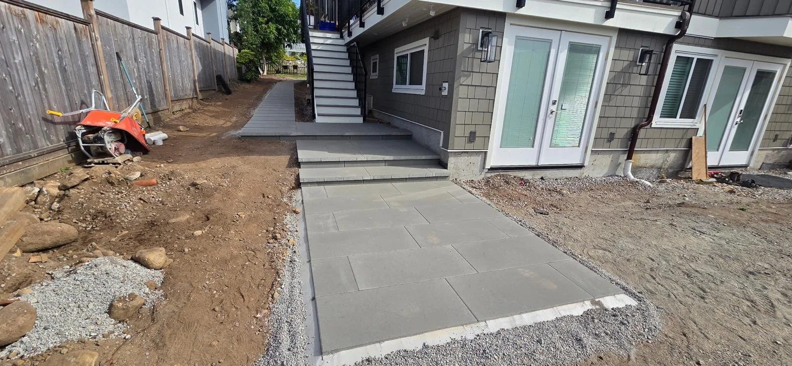 Newly paved concrete walkway leading to back door of a house under construction, with stairs, and a neighboring fenced yard on the left.