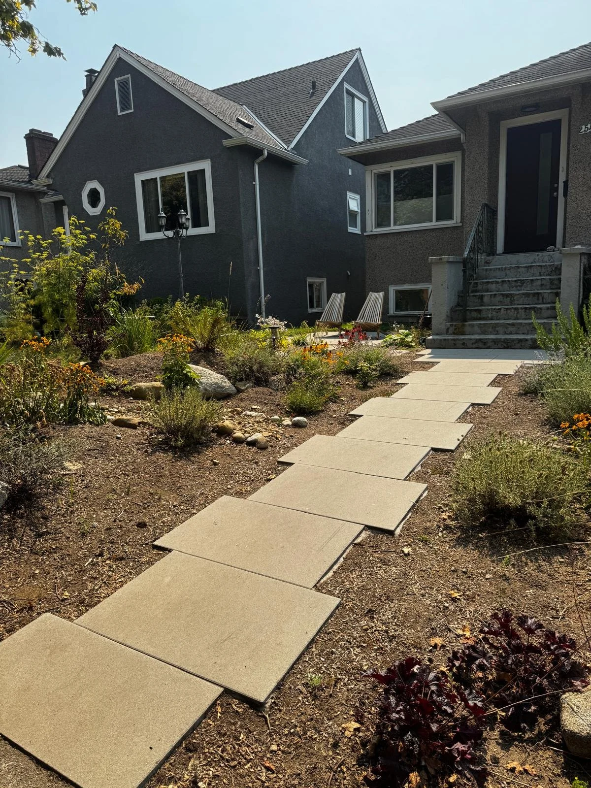 A backyard with a paved walkway leading to house steps, surrounded by garden plants and flowers, with neighboring houses in the background.