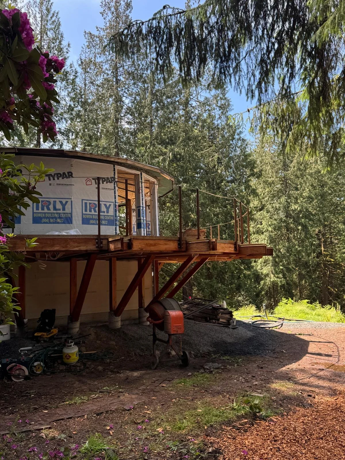 House under construction with a wooden deck, surrounded by trees and bushes, construction tools and materials on the ground.