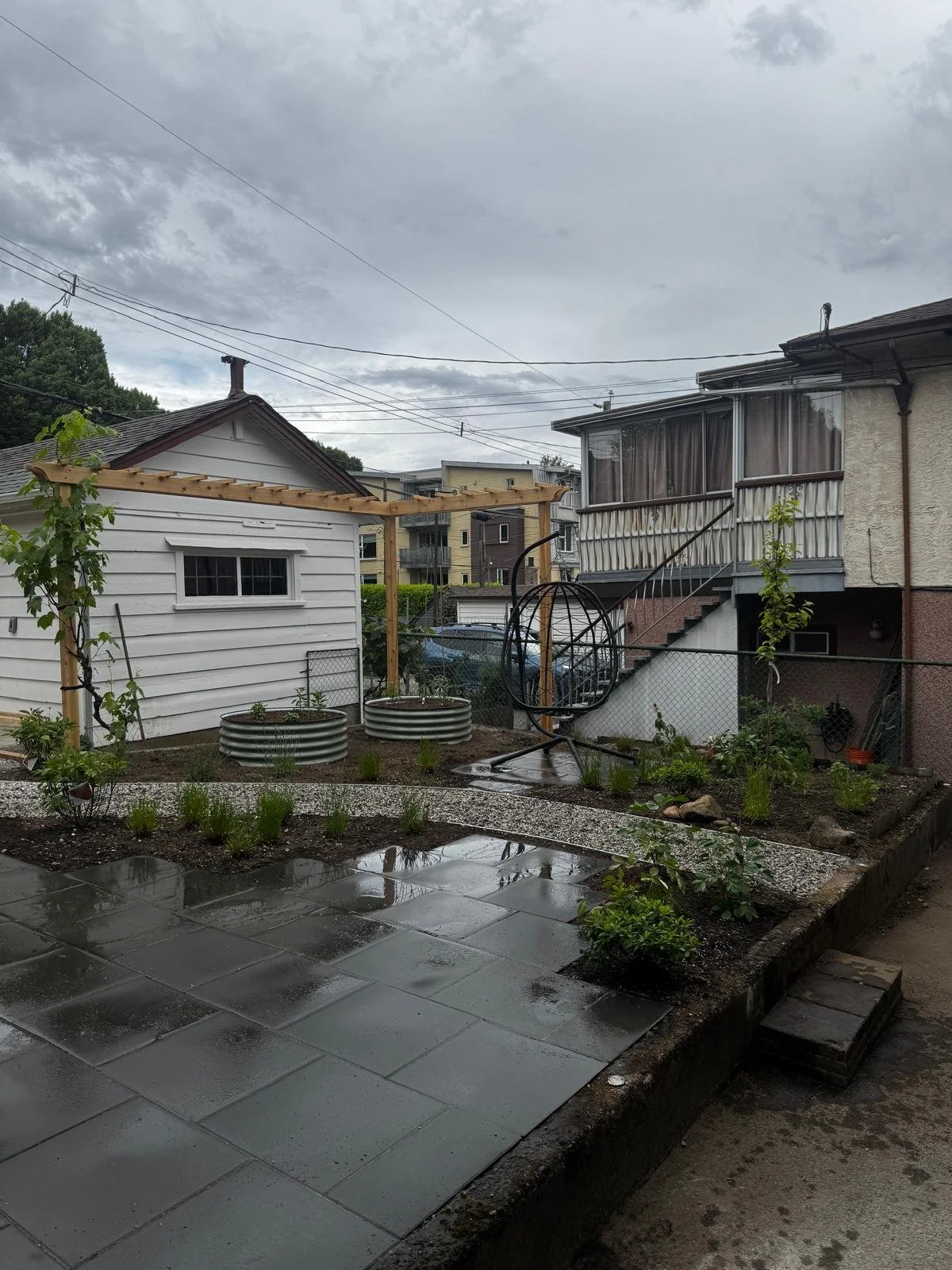 Backyard garden with newly installed gray paver patio, potted plants, and garden beds under an overcast sky.