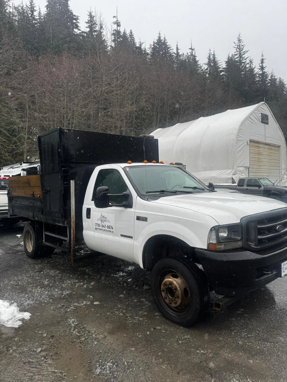 A white work truck parked outdoors with a black cargo bed, in front of a large white tent and a forested hillside, on a cloudy day with patches of snow on the ground.