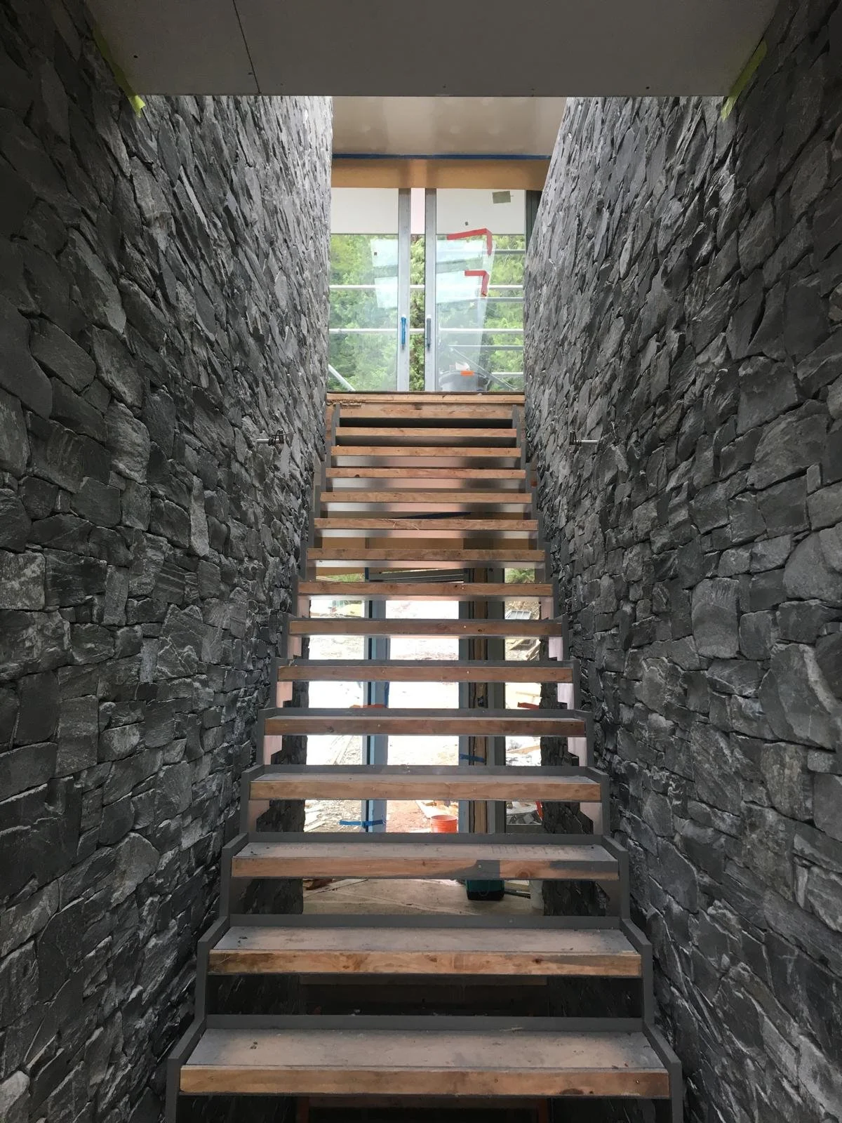 Interior view of a building under construction, showing a staircase with wooden steps, stone walls on both sides, and a glass door at the top leading outside.