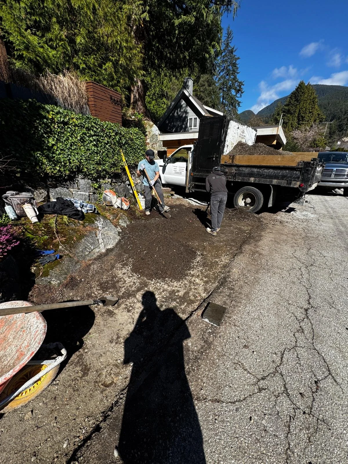 Workers repairing a damaged road surface next to a grassy hillside with trees, a small house, and a mountain in the background. They are using shovels and a wheelbarrow.