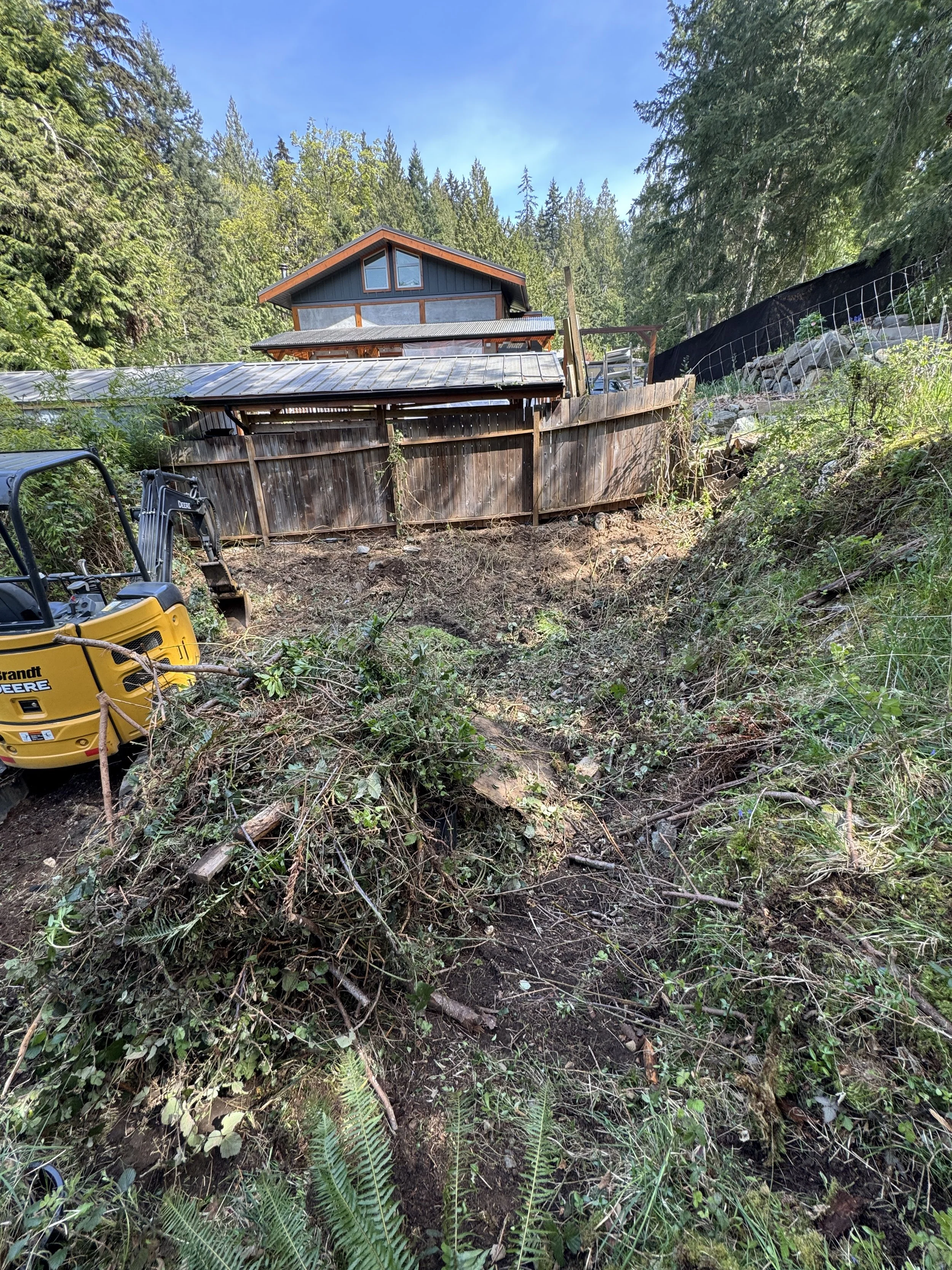 A construction site with a yellow mini excavator and a pile of dirt and branches in the foreground. In the background, a wooden fence, a house with a dark blue exterior and large windows, surrounded by trees under a partly cloudy blue sky.
