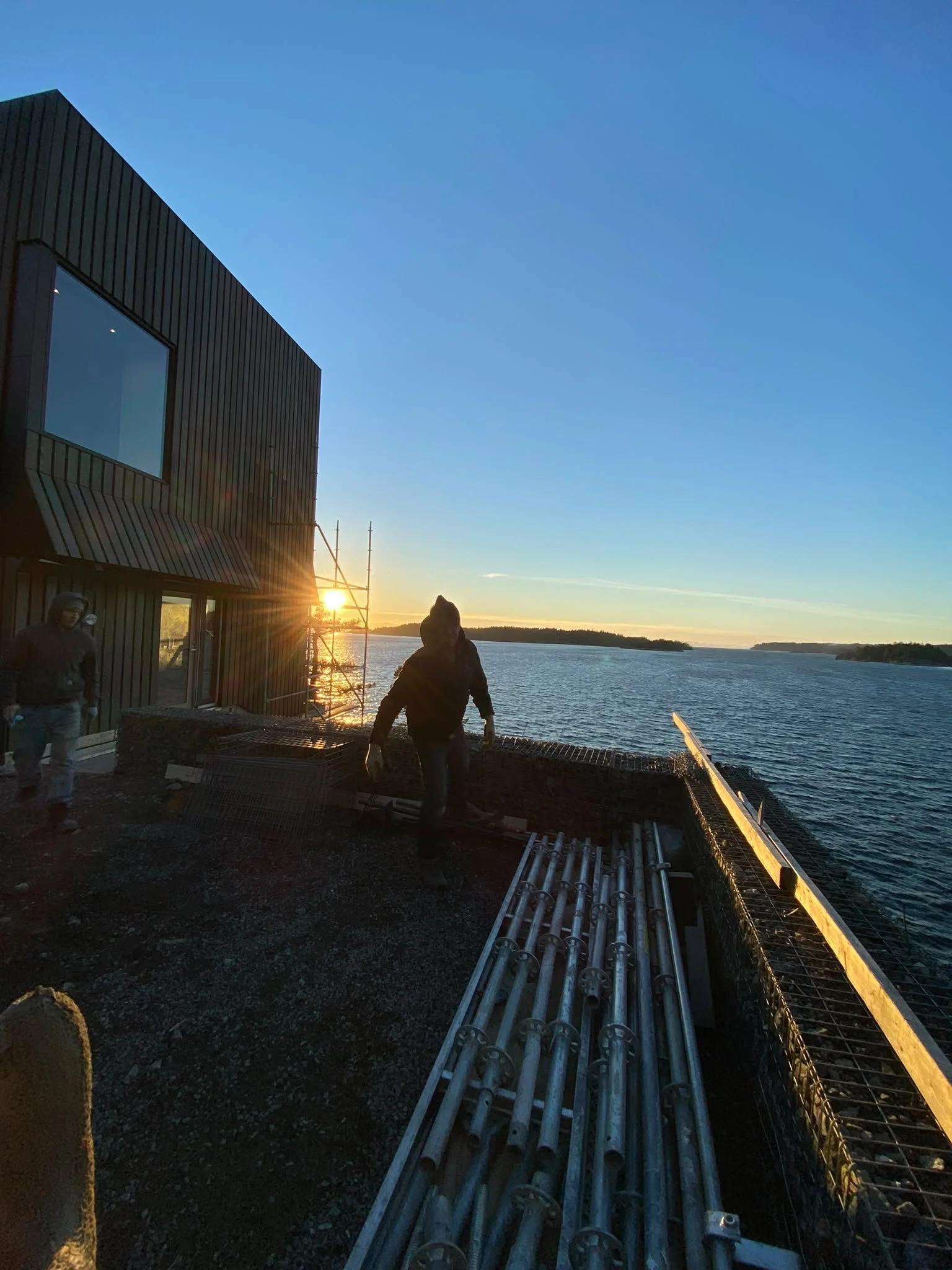 Construction workers installing metal pipes on a building by water during sunset, with clear sky and distant land.