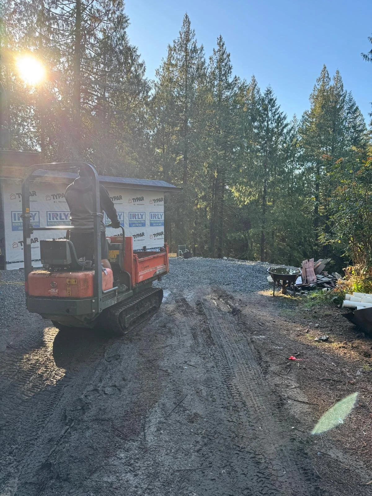 A construction site in a forested area during daylight with a small tracked excavator and a partially built structure covered in Typar weather-resistant barrier, and some construction materials including a wheelbarrow and pipes on the right side.