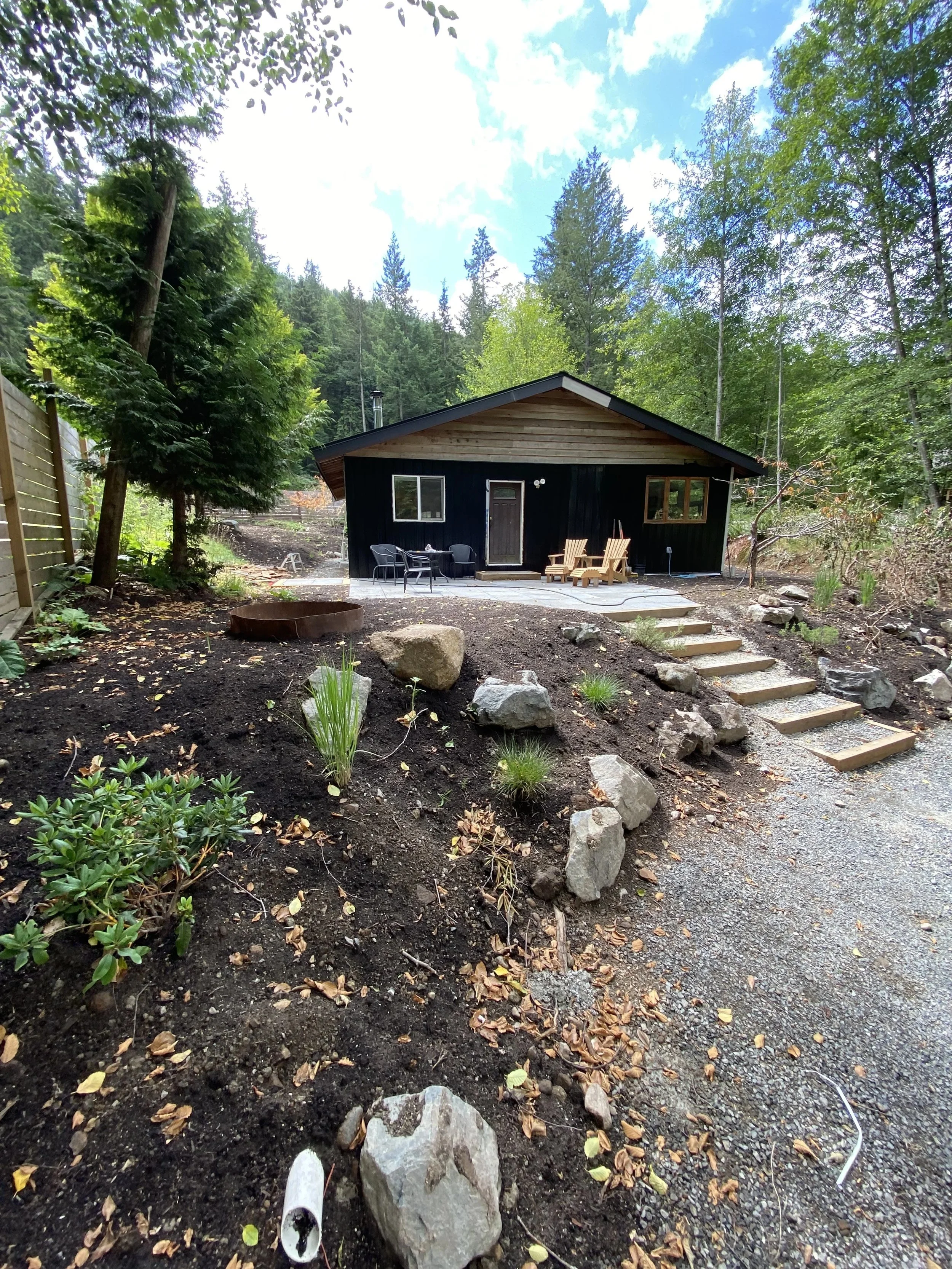 Backyard with a small black house, outdoor furniture, and natural landscaping surrounded by trees and mountains under a partly cloudy sky.