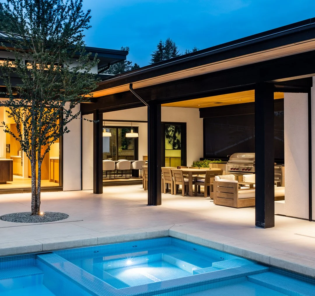 A modern house outdoor patio at dusk with a lit swimming pool, wooden dining table with chairs, a built-in barbecue grill, and interior lighting visible through large windows.