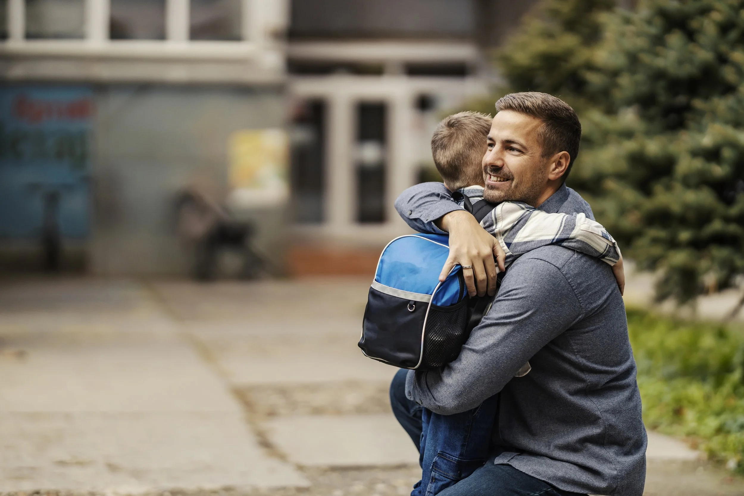 Man smiling and hugging a child outdoors, holding a backpack, with a blurred urban background.