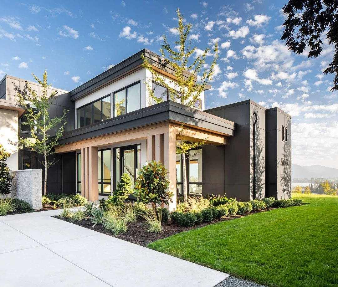Modern black and beige house with large windows, landscaped yard, and blue sky with clouds.