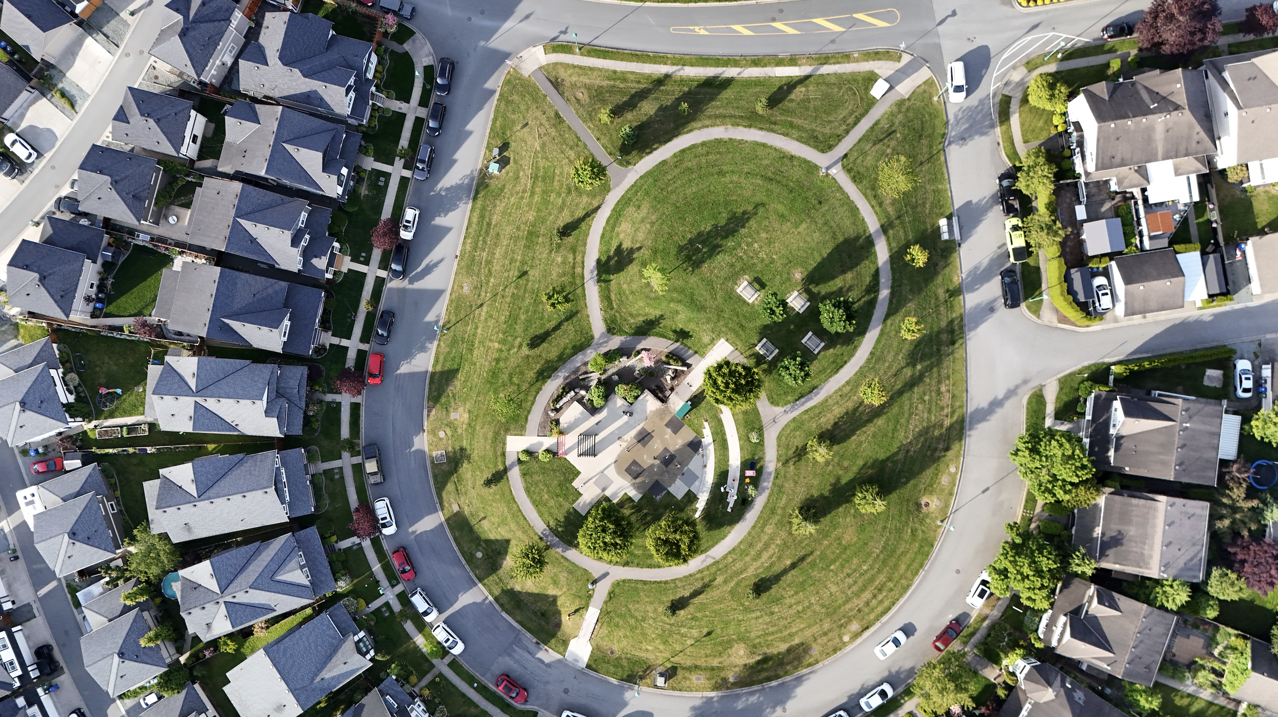 Aerial view of a neighborhood park with green lawns, trees, benches, pathways, and a circular plaza, surrounded by houses, streets, and parked cars.