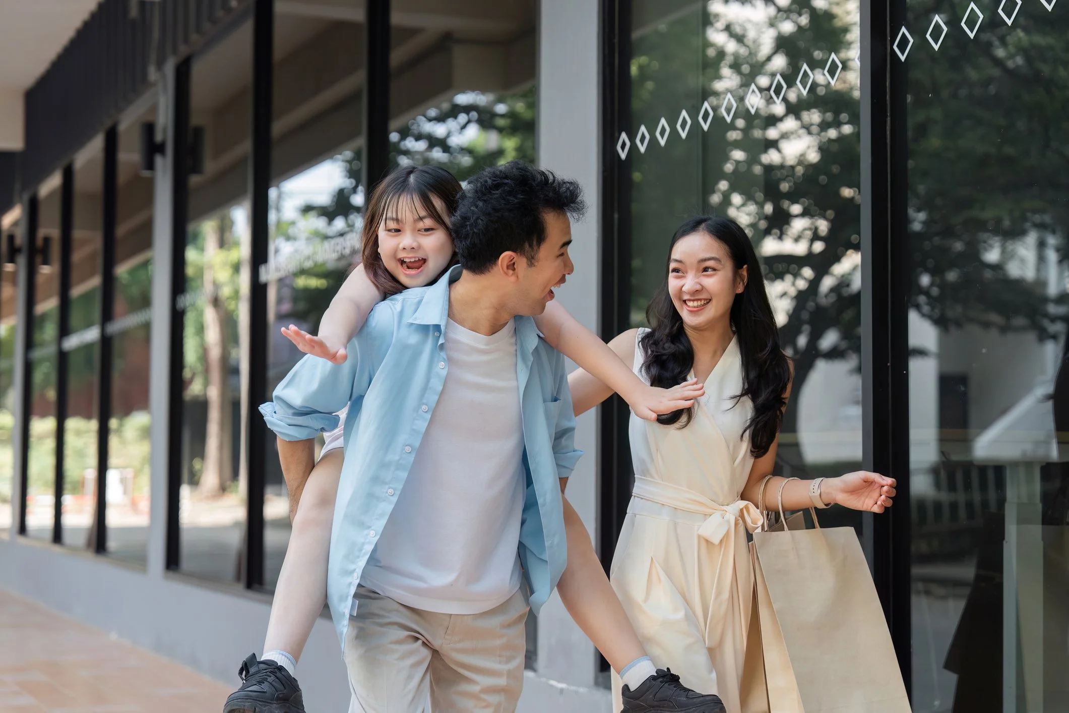 A group of three people, two women and a man, smiling and laughing outside a modern building, with the man giving a piggyback ride to a young girl who is also smiling and appears happy.