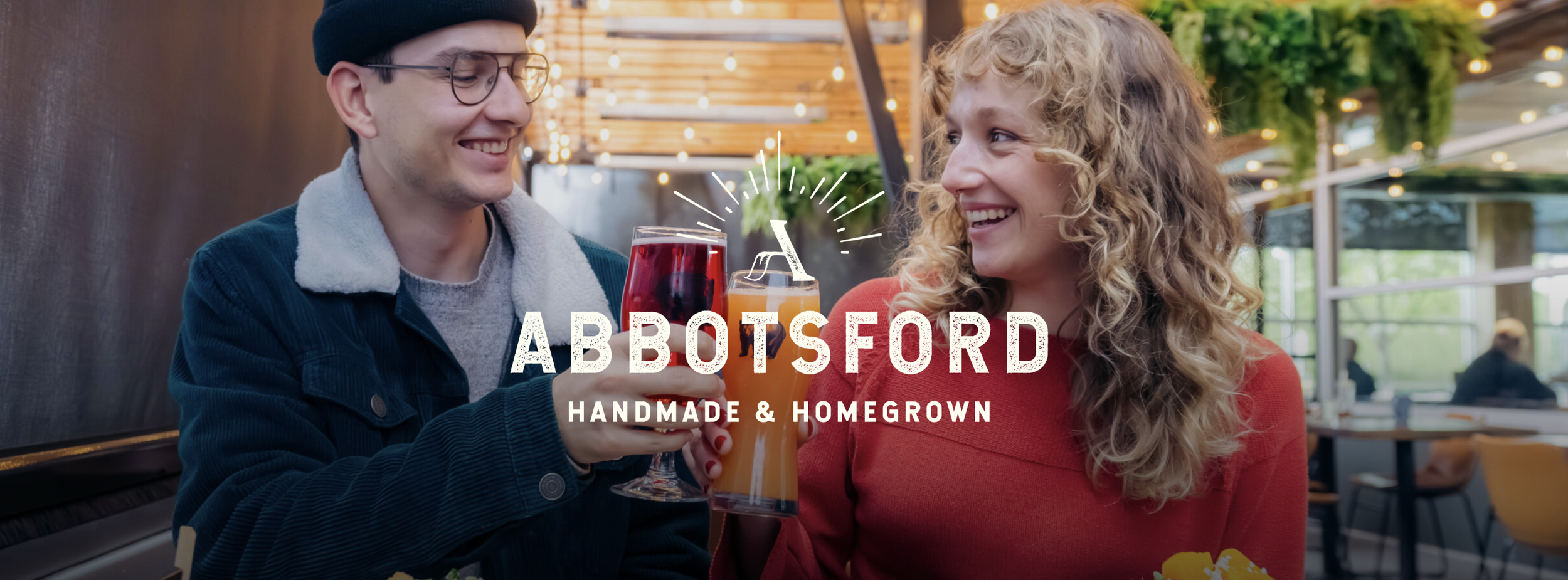 A man and woman smiling and holding drinks at a restaurant with a logo for Abbotsford Handmade & Homegrown overlayed on the image.