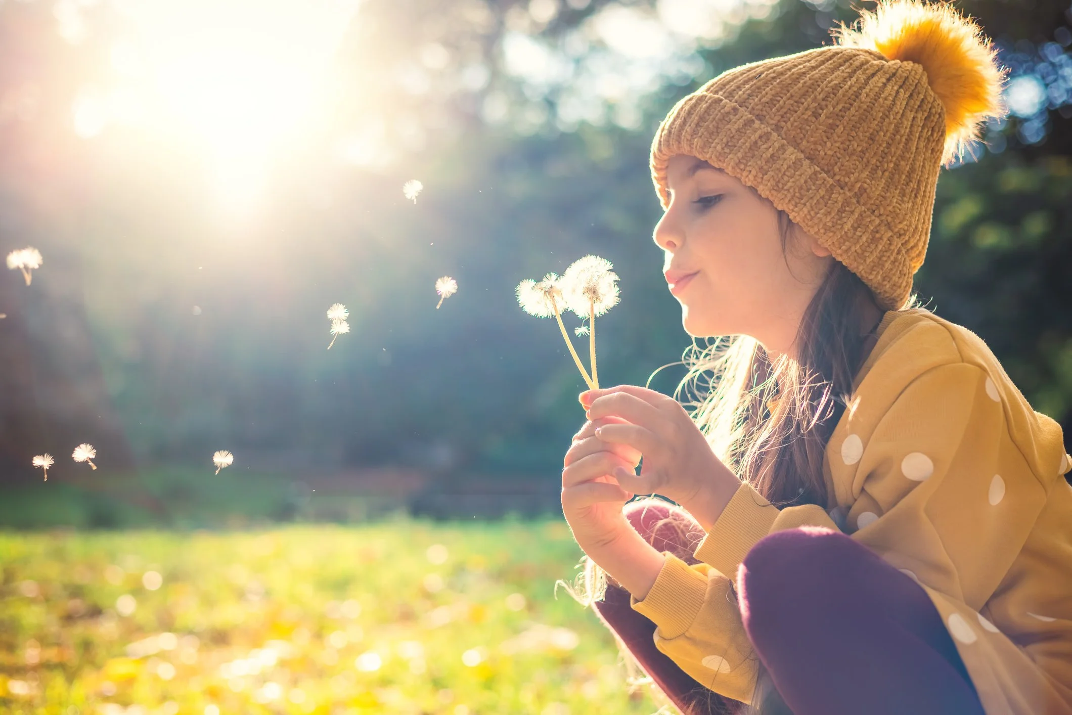 A young girl wearing a mustard-colored beanie with a pom-pom, a yellow jacket, and purple pants, kneeling on the grass while blowing on dandelions in a park during golden hour.