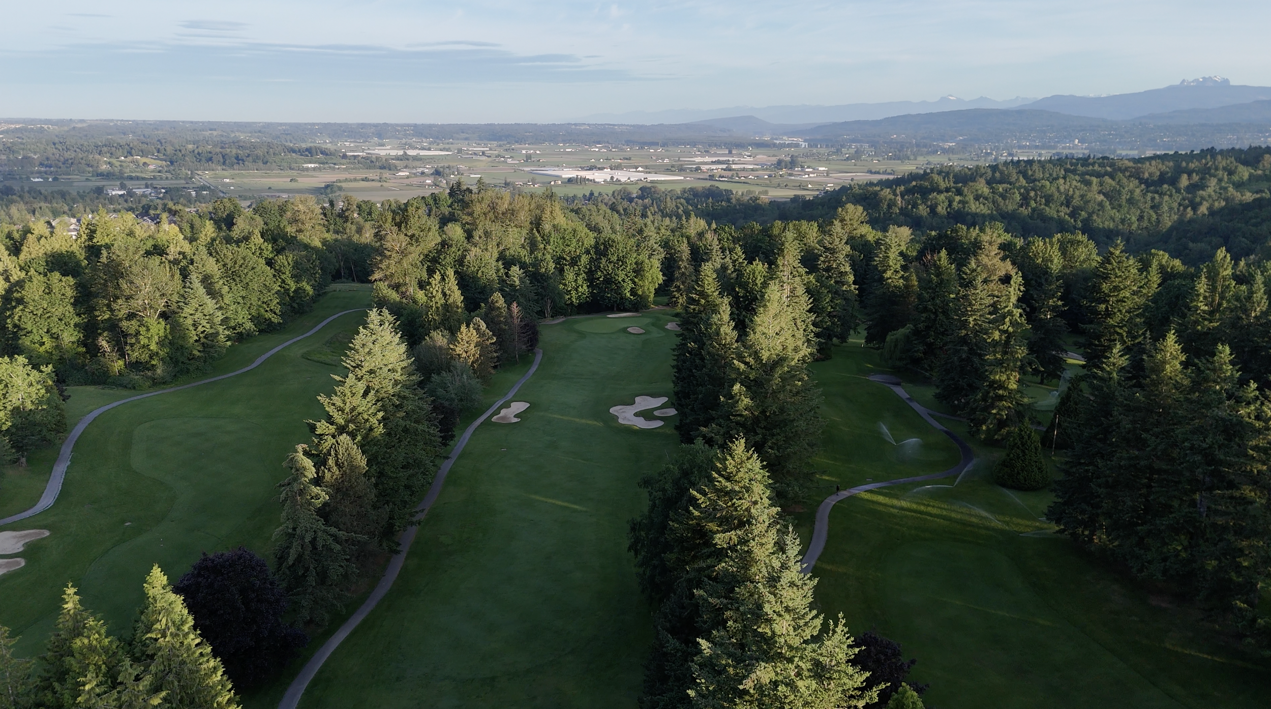Aerial view of a lush green golf course surrounded by trees with a distant cityscape and mountains in the background.
