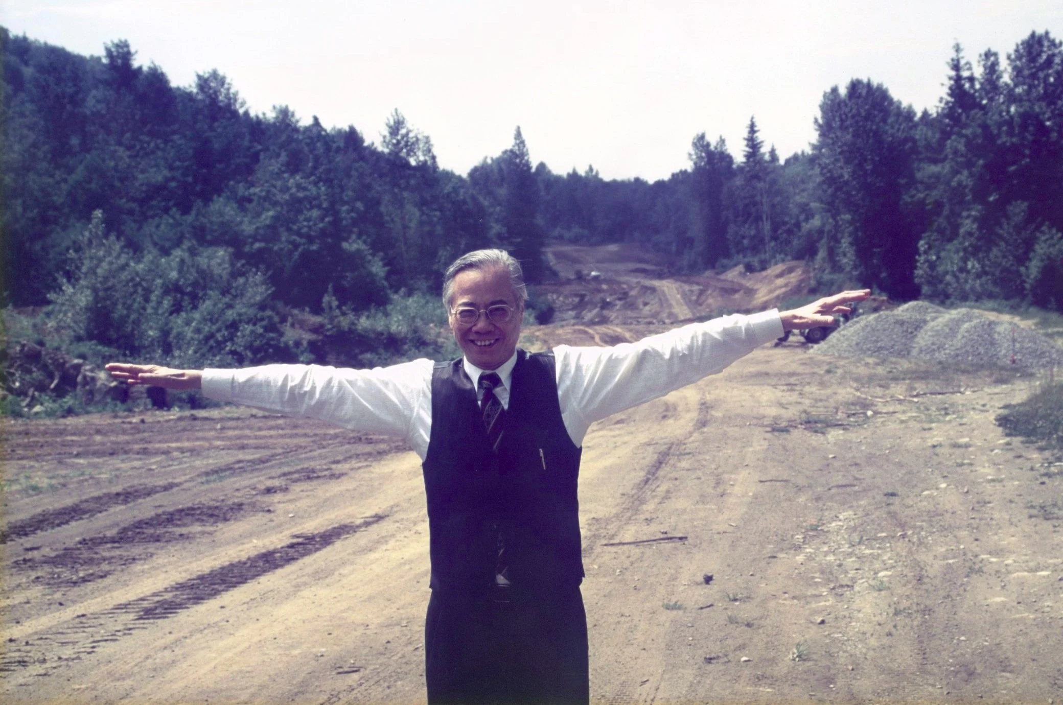 A man in formal attire with glasses, smiling, and extending his arms outward outside on a dirt road with wooded hills in the background.