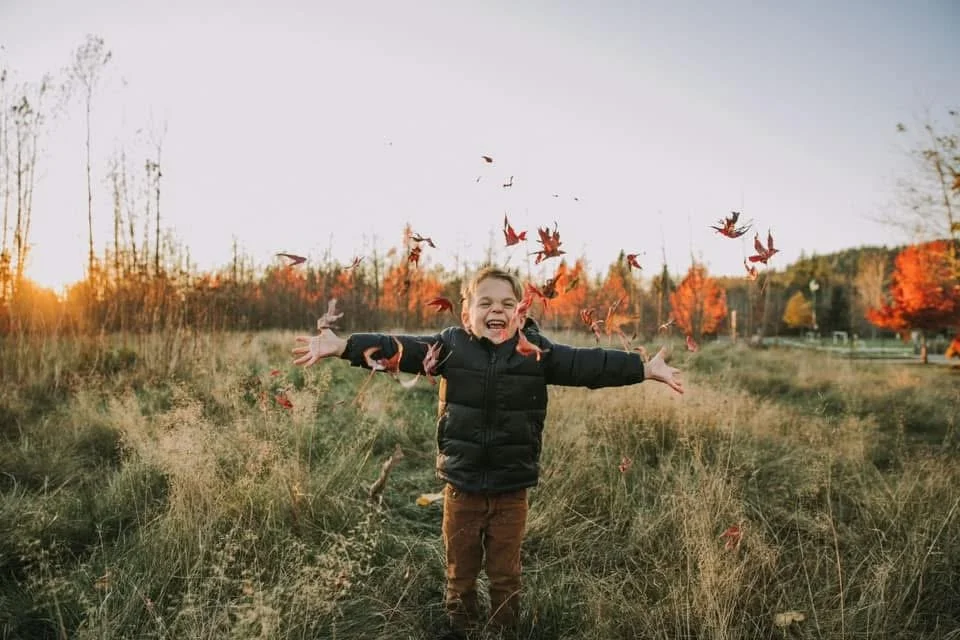 Child with arms outstretched joyfully in a field during autumn, surrounded by falling red and orange leaves with trees in the background.