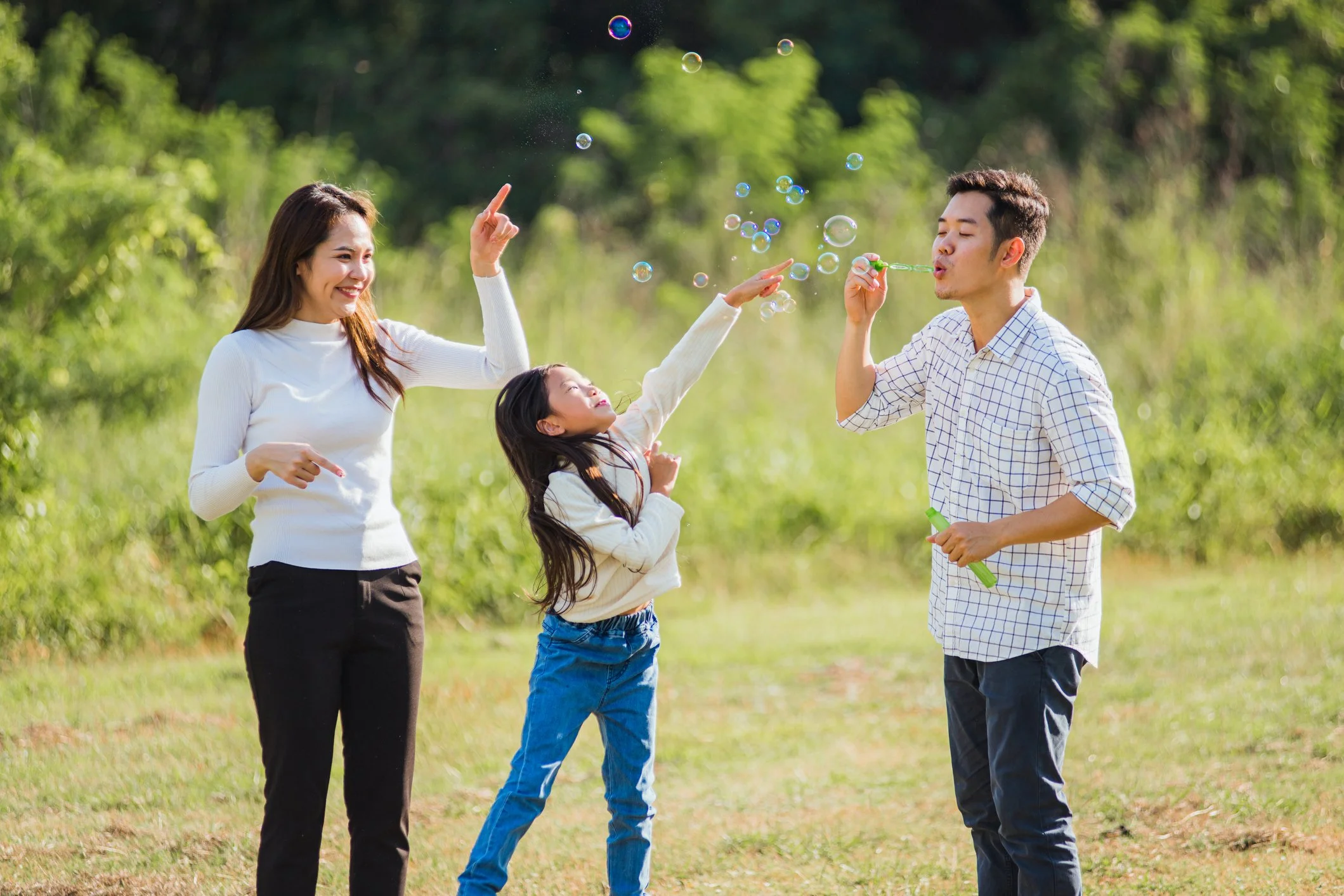A family of three enjoying outdoor activities on a sunny day, blowing bubbles in a park with green trees and grass.