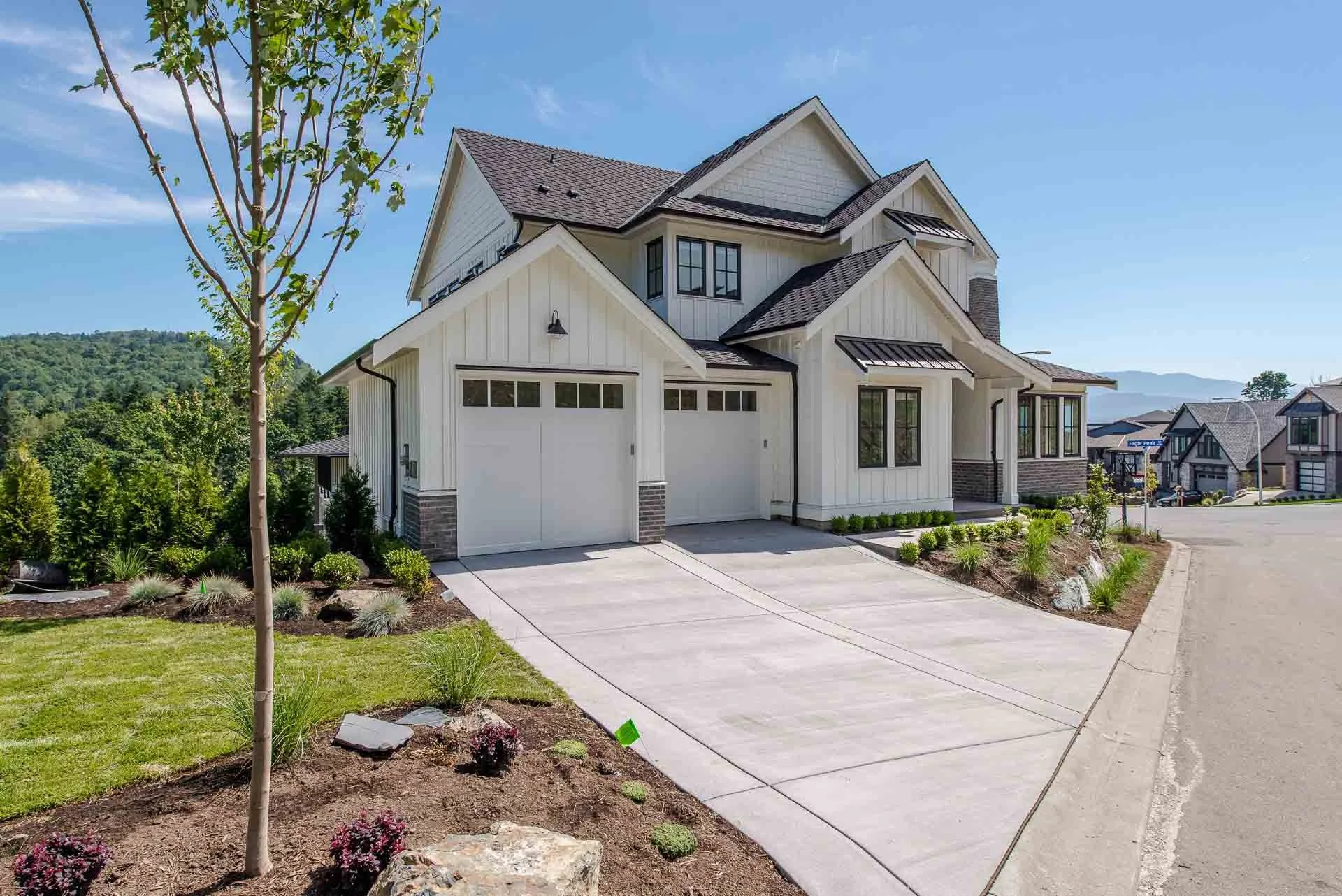 A modern two-story house with a white exterior, dark roofing, and a garage with white doors. The front yard features a garden with green plants, small trees, and mulch, with a concrete driveway leading to the garage. There are neighboring houses visible in the background and a clear blue sky.