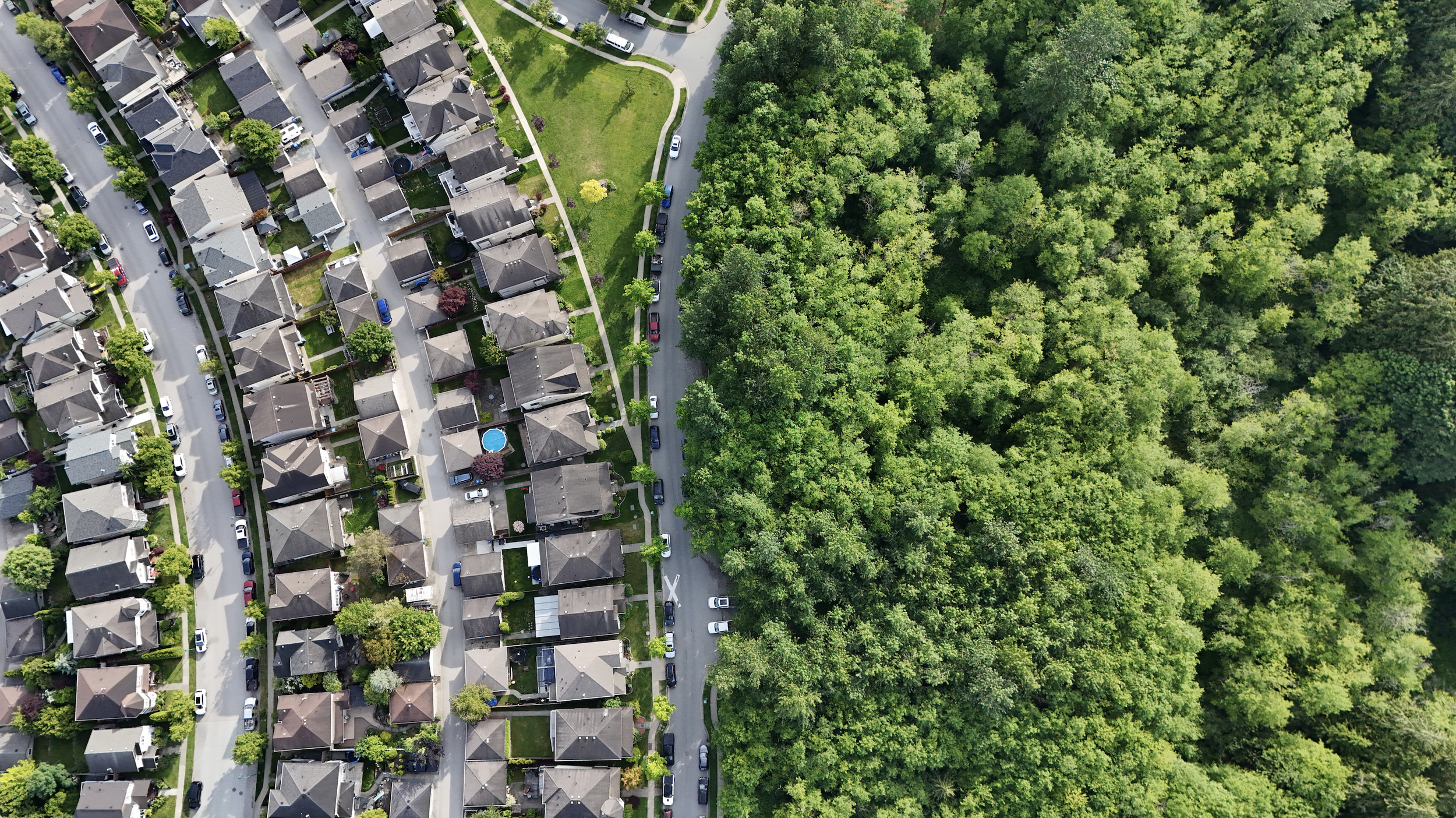 An aerial view of a neighborhood with houses, trees, and a large wooded area on the right side.