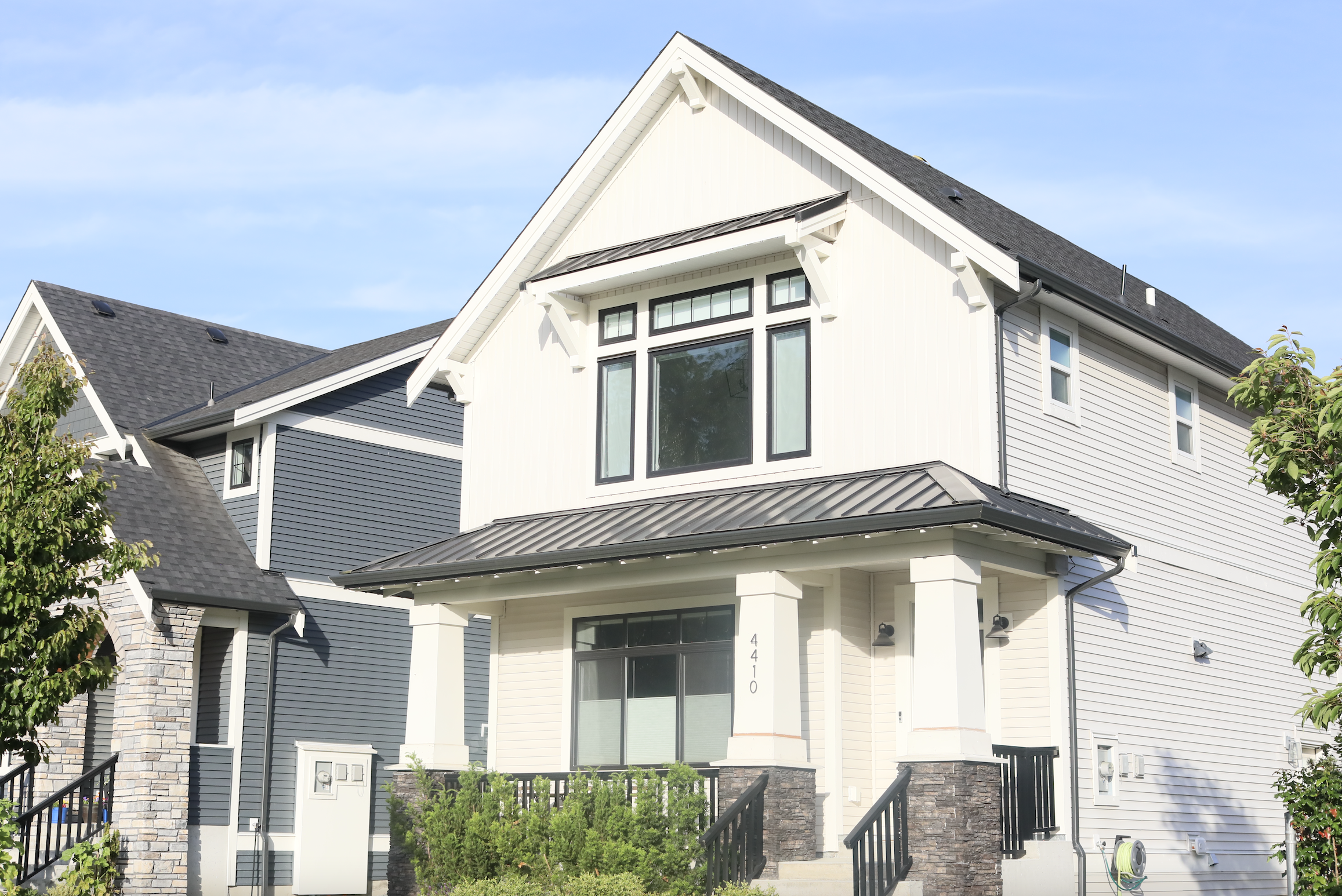 Modern multi-story house with white siding, gray accents, large front windows, and a steep gabled roof, surrounded by greenery and a blue sky.