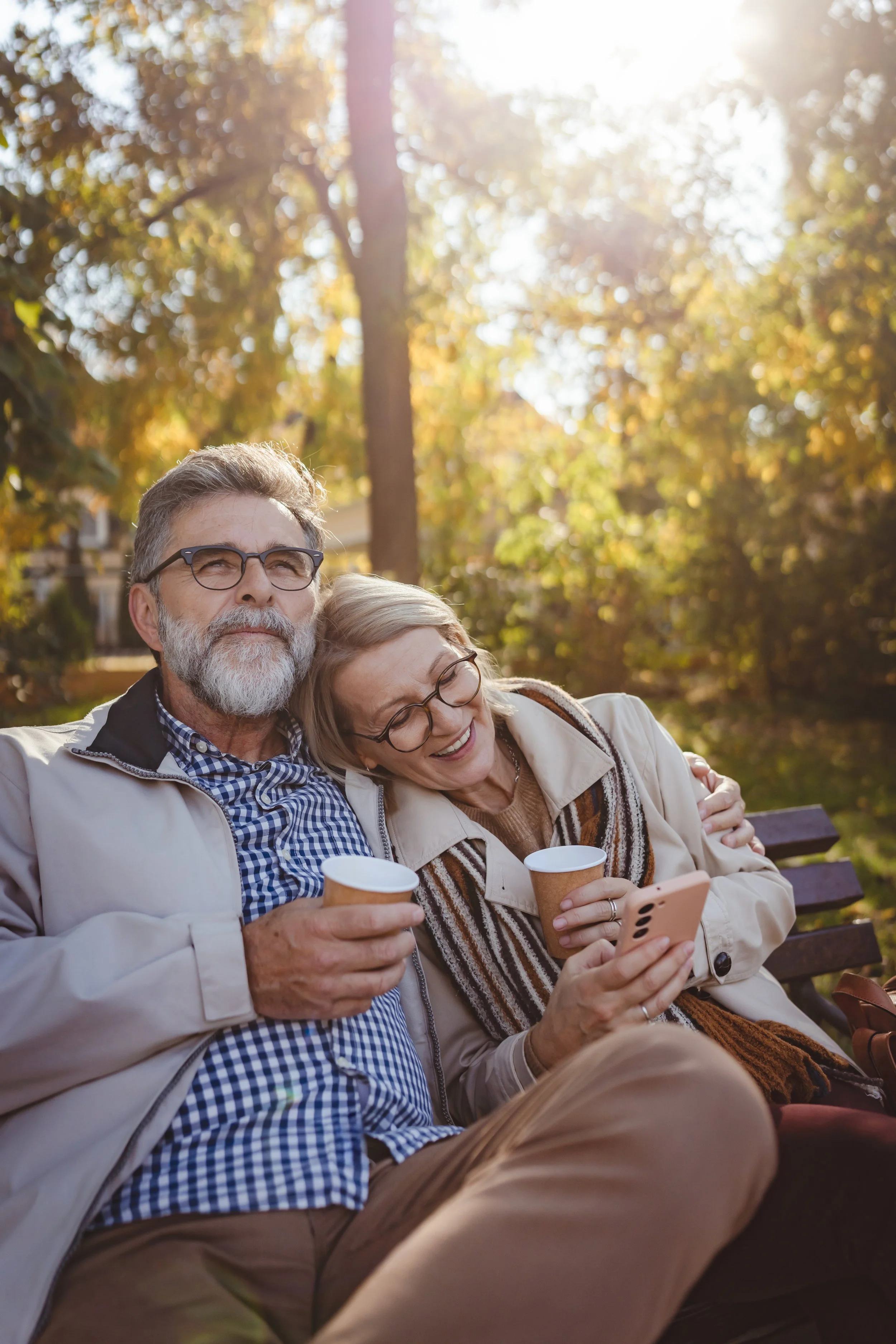 An elderly couple sitting on a park bench, smiling and looking at a smartphone. They are holding coffee cups. The background has trees with autumn leaves and sunlight.