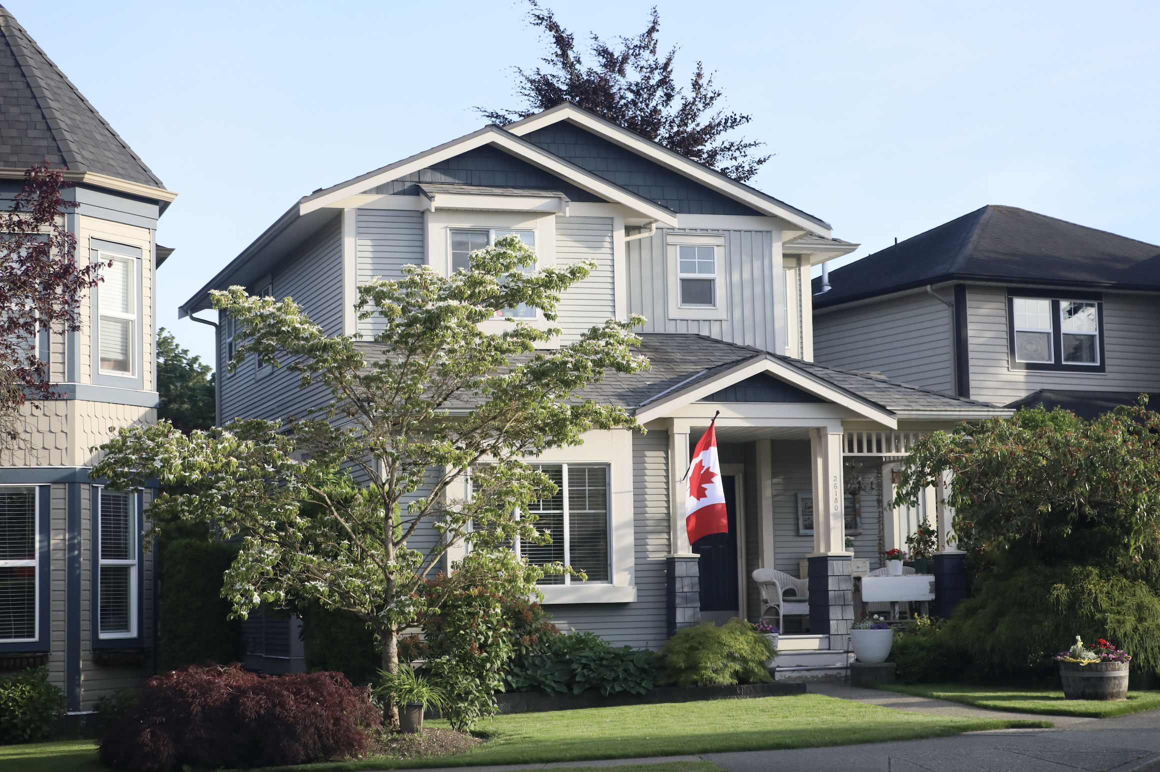 A large, gray, two-story house with white trim, surrounded by greenery and trees, with a Canadian flag displayed near the front porch.