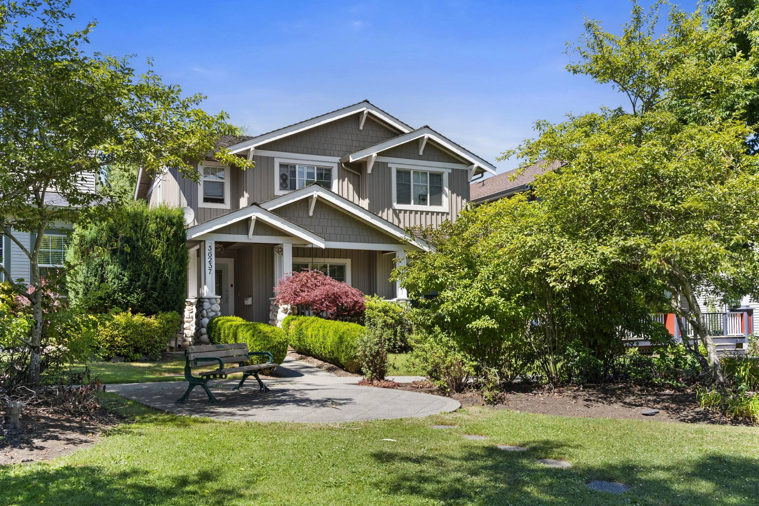 A multi-story house with gray siding, white trim, and a front porch, surrounded by lush green trees and bushes, with a bench on a paved pathway in the front yard on a bright, clear day.