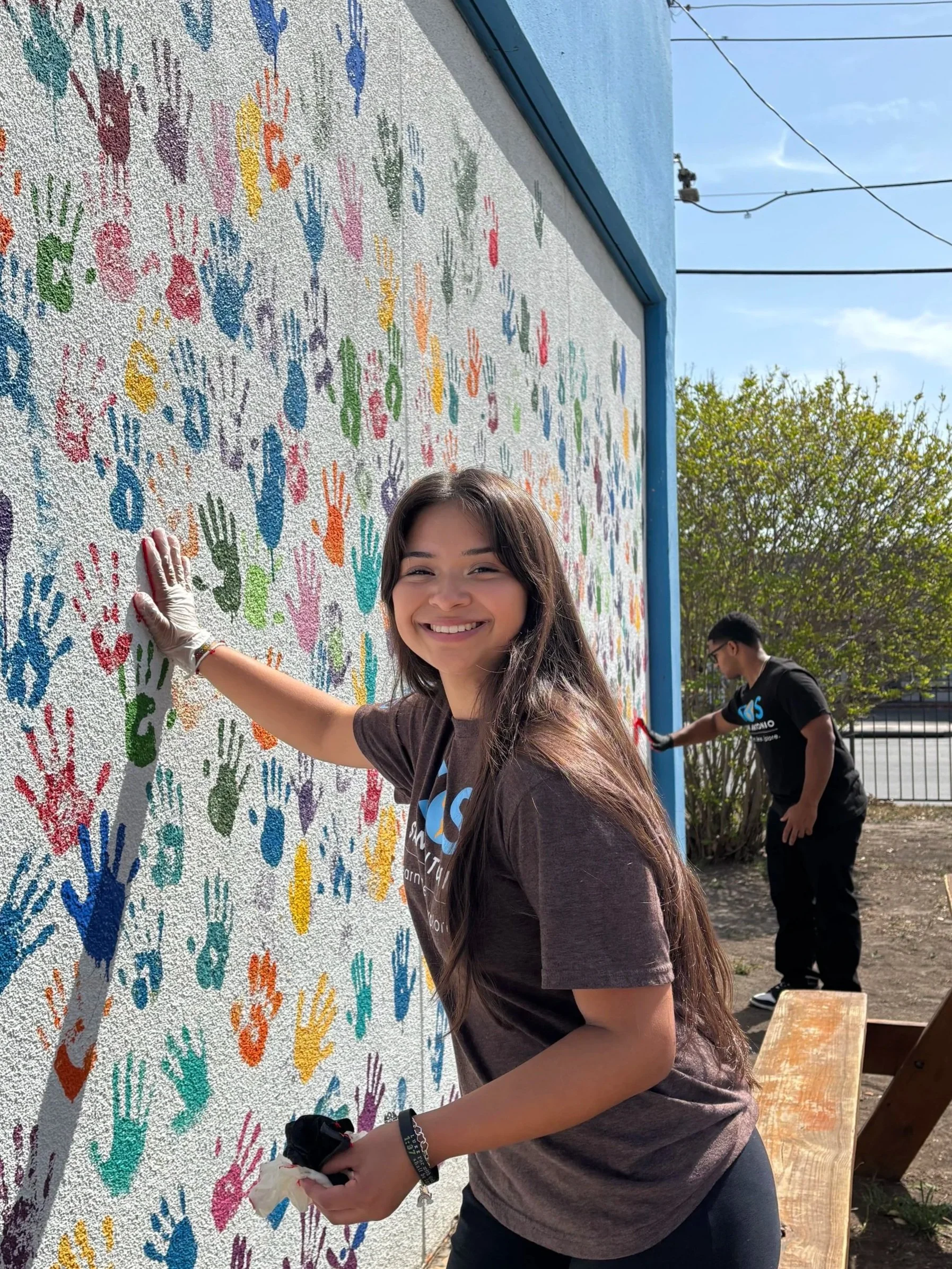 Young woman with long brown hair smiling as she paints colorful handprints on a wall with a mural, while a person in the background also painting on the wall.