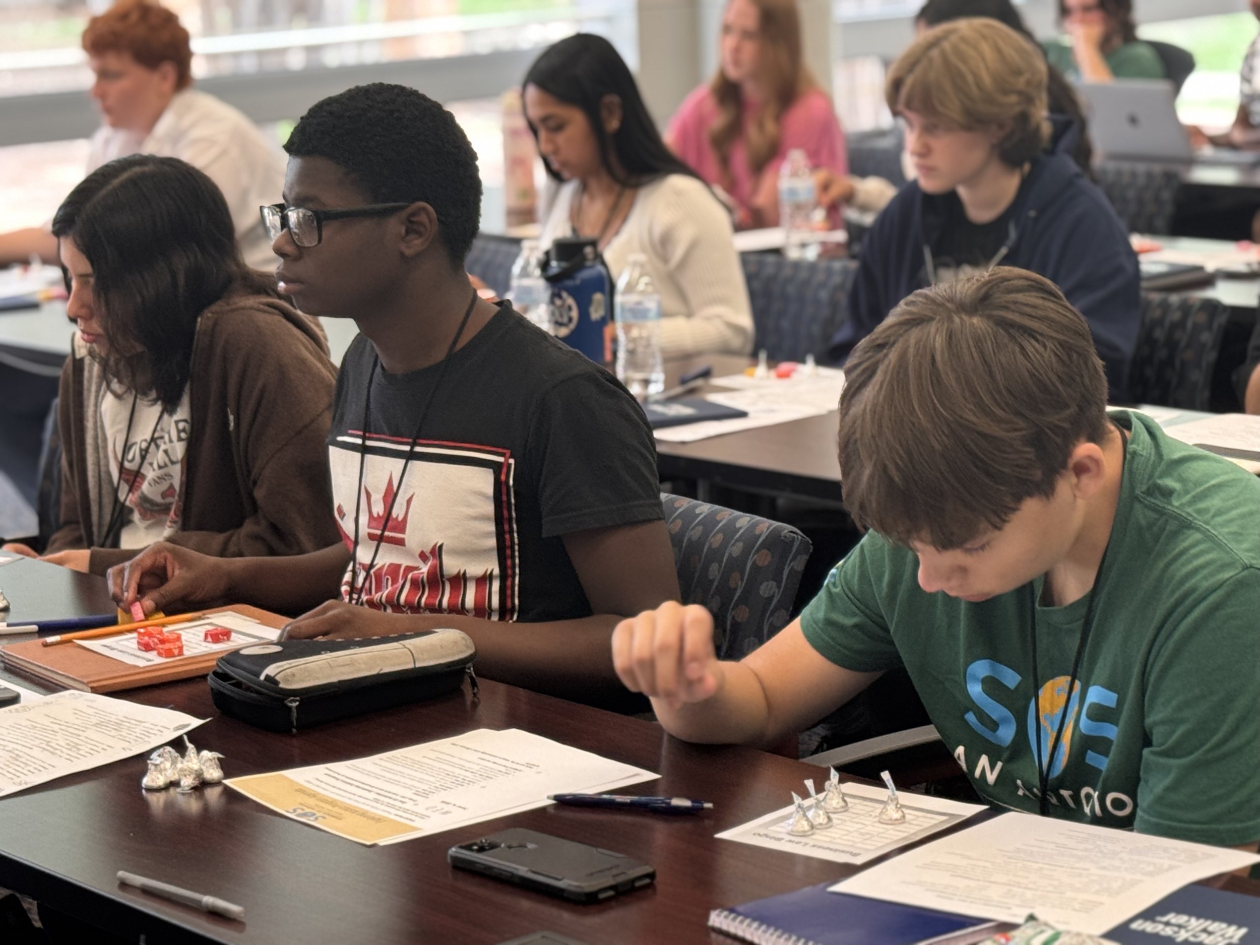 Students sitting at a classroom desk, engaged in activities with papers, markers, and game pieces, in a well-lit room.