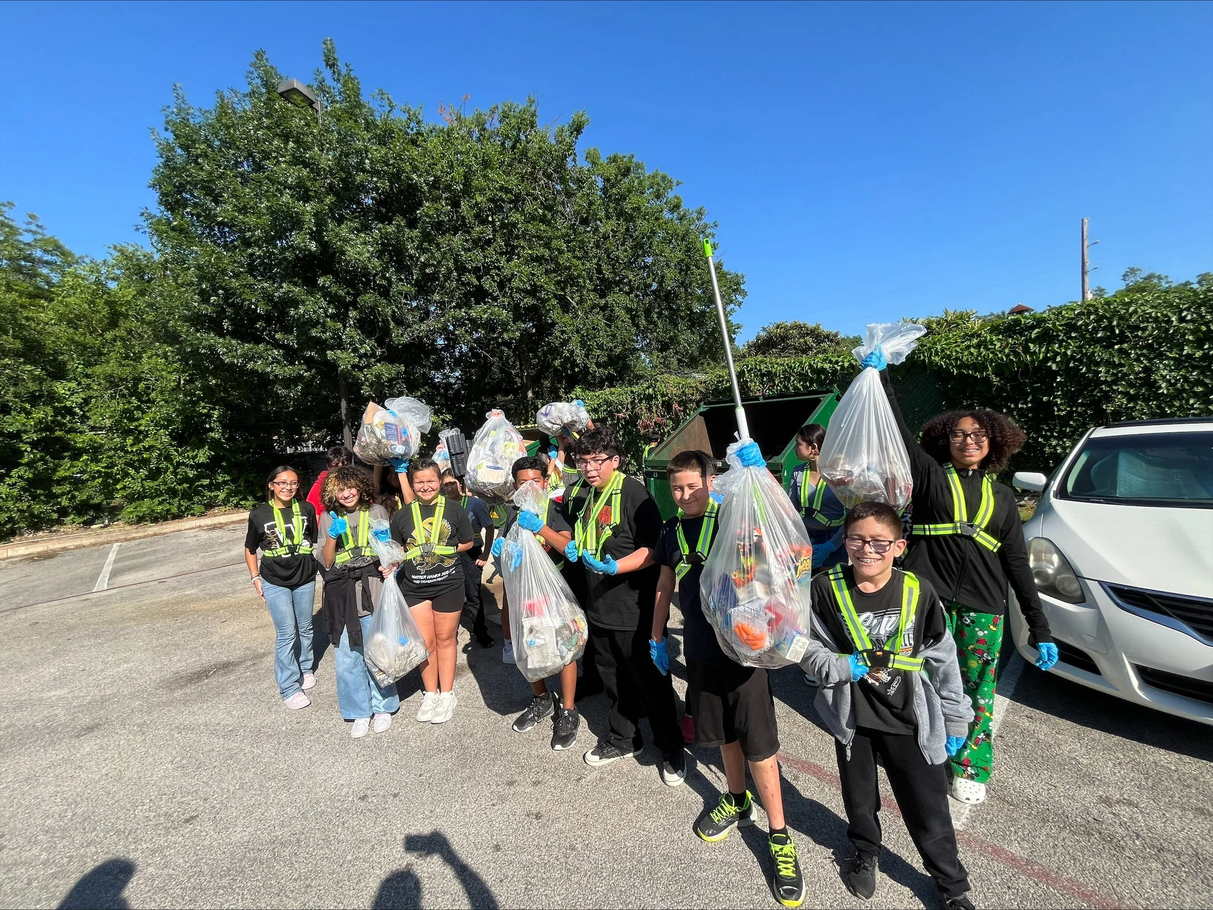 Group of children and teenagers participating in a community cleanup. They are holding large garbage bags filled with trash and wearing gloves. The event is taking place outdoors on a sunny day, with trees and a parked car in the background.