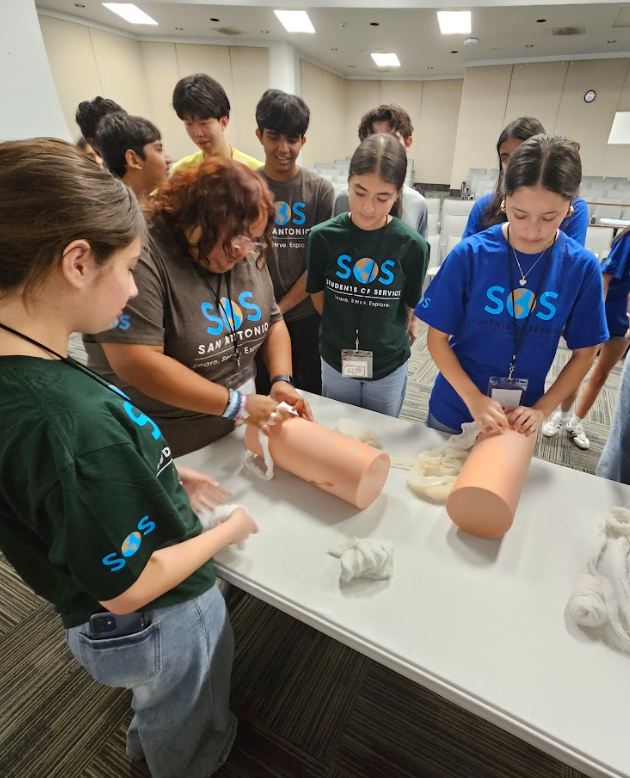 Group of children and an adult practicing bandaging on a plastic arm model in a classroom setting.