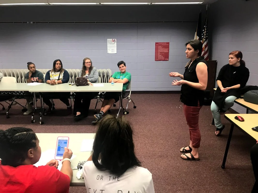 A woman is speaking to a small group of students in a classroom. The students are seated at tables while the woman stands in front of them, gesturing as she talks. The room has a purple walls and some posters.