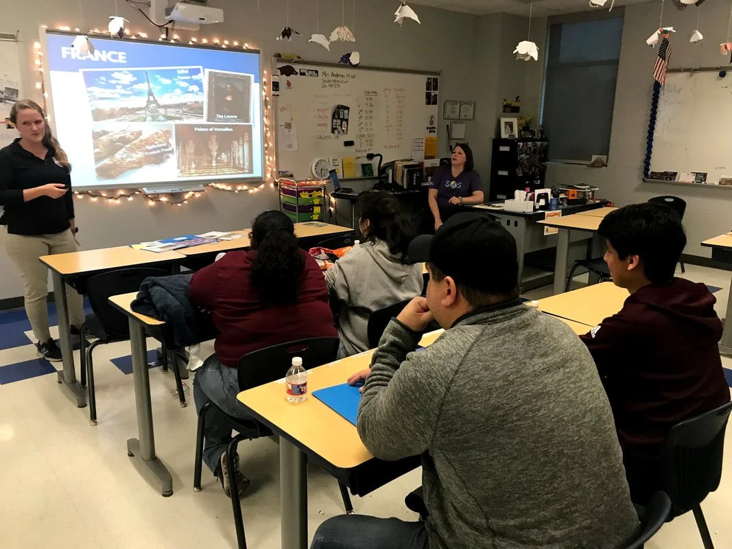A classroom with students seated at desks, listening to a teacher giving a presentation about international travel.