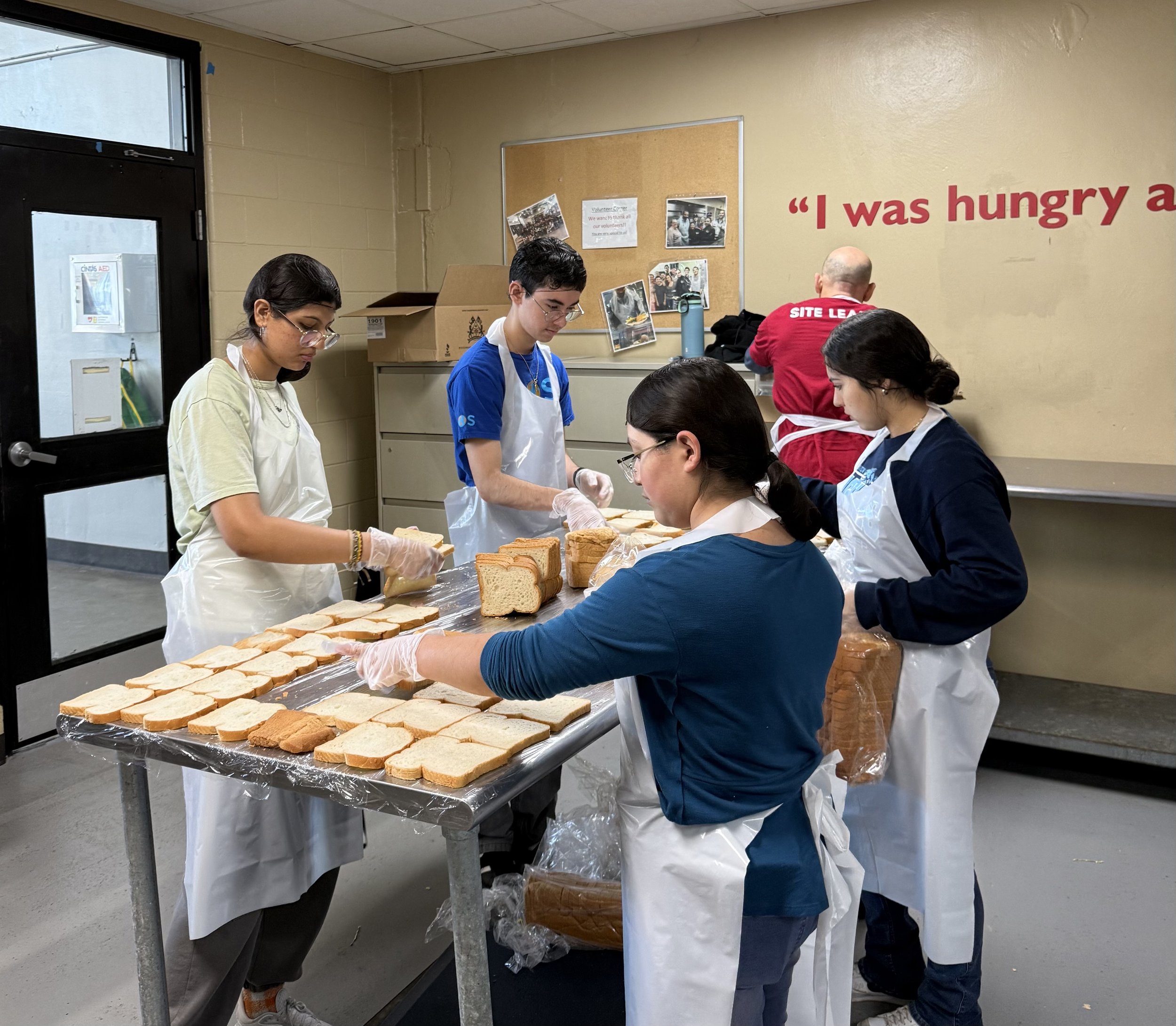 Five people wearing aprons and gloves are packing slices of bread on a table in a food packaging kitchen. Others are in the background preparing more bread. The room has a beige wall with the quote 'I was hungry a' partially visible.