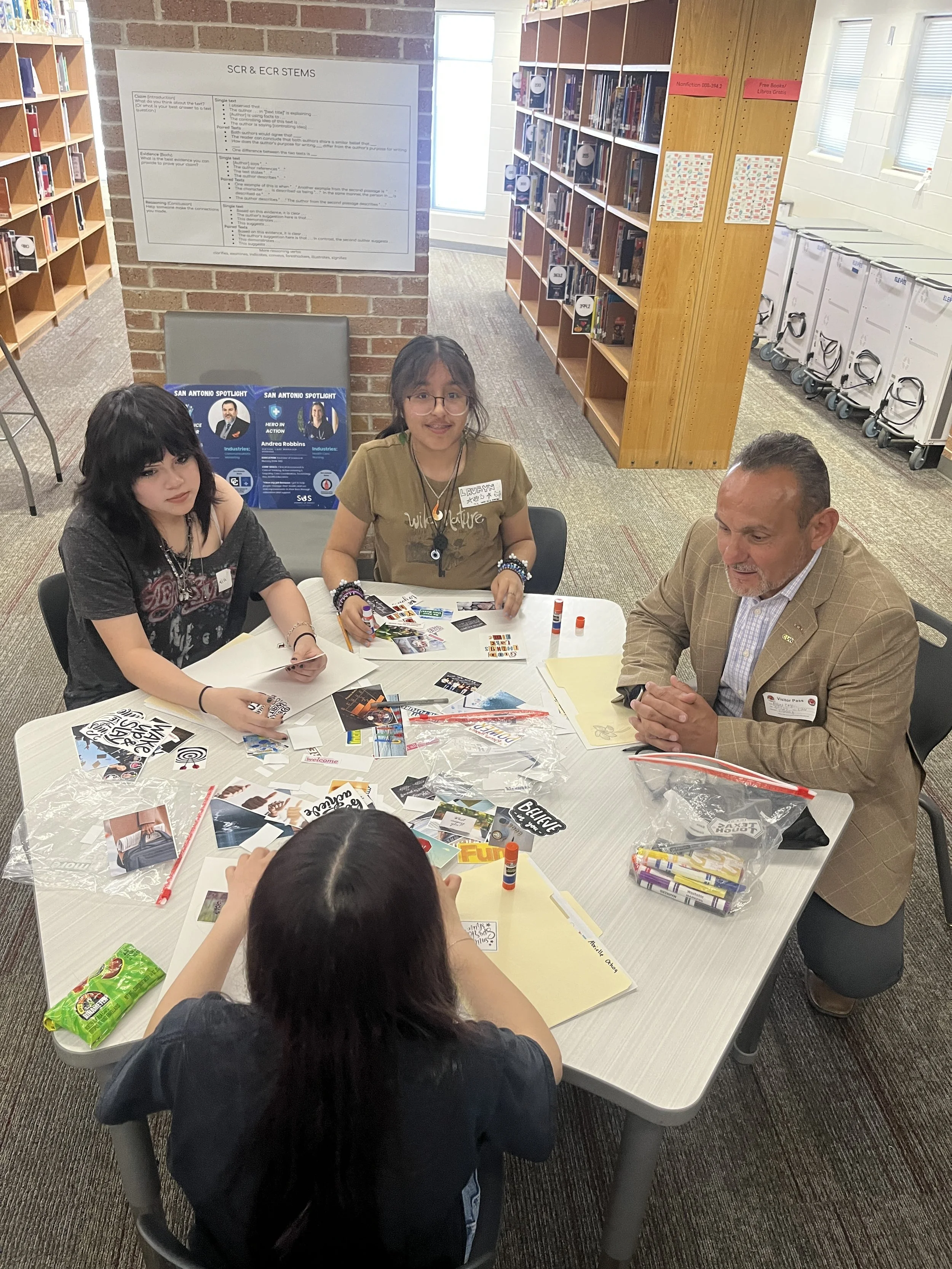 A group of children and an adult sitting at a table in a library, engaging in a creative activity with various craft supplies and photo cards spread on the table.