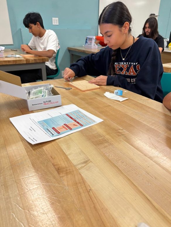 Students in a classroom practicing with medical or first aid training supplies, including a practice kit and instruction sheet on the wooden table.