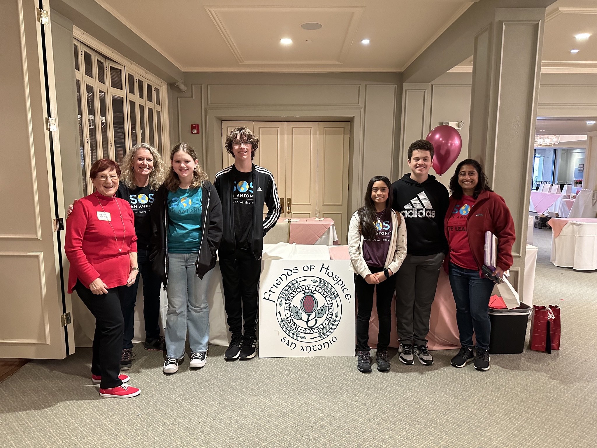 Group of eight people standing indoors behind a table with a Friends of Hospice logo, some wearing SOS San Antonio T-shirts, in a banquet hall setting with pink tablecloths and a pink balloon.