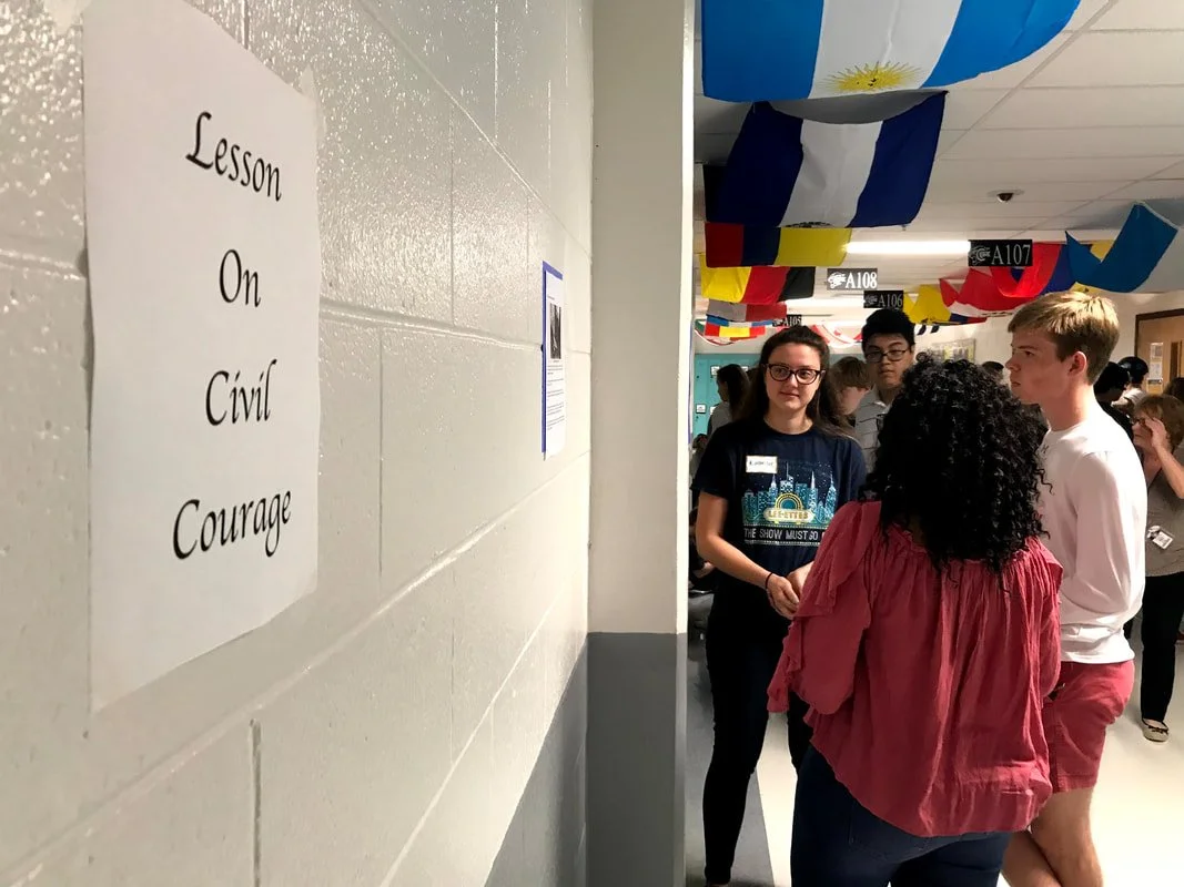 A hallway in a school decorated with flags, with students standing and talking near classroom doors, and a sign on the wall that reads 'Lesson on Civil Courage'.