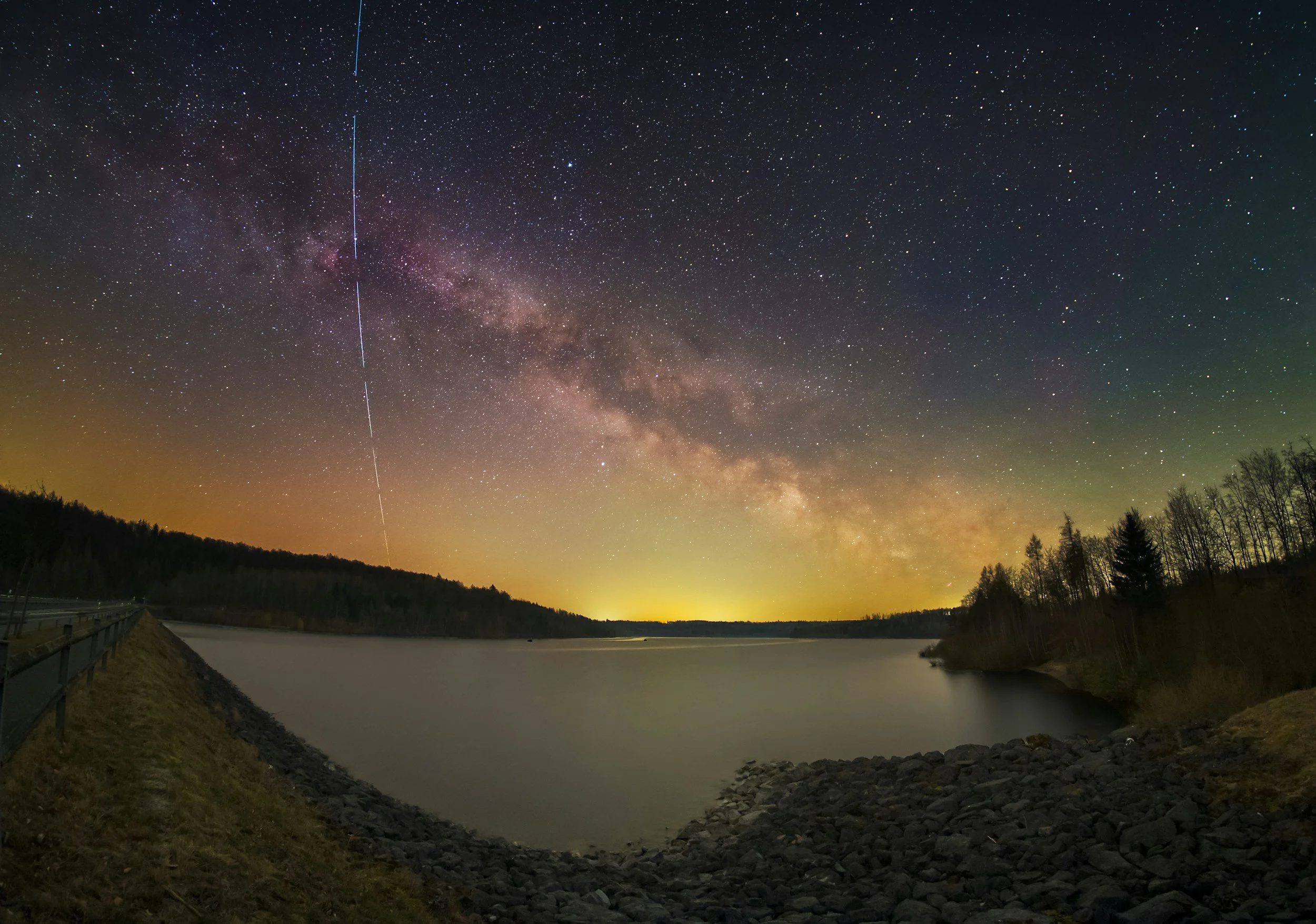 Nighttime scene of a lake with a rocky shore and forested hills in the background. The sky is filled with stars, the Milky Way galaxy, and streaks of light from the sky.