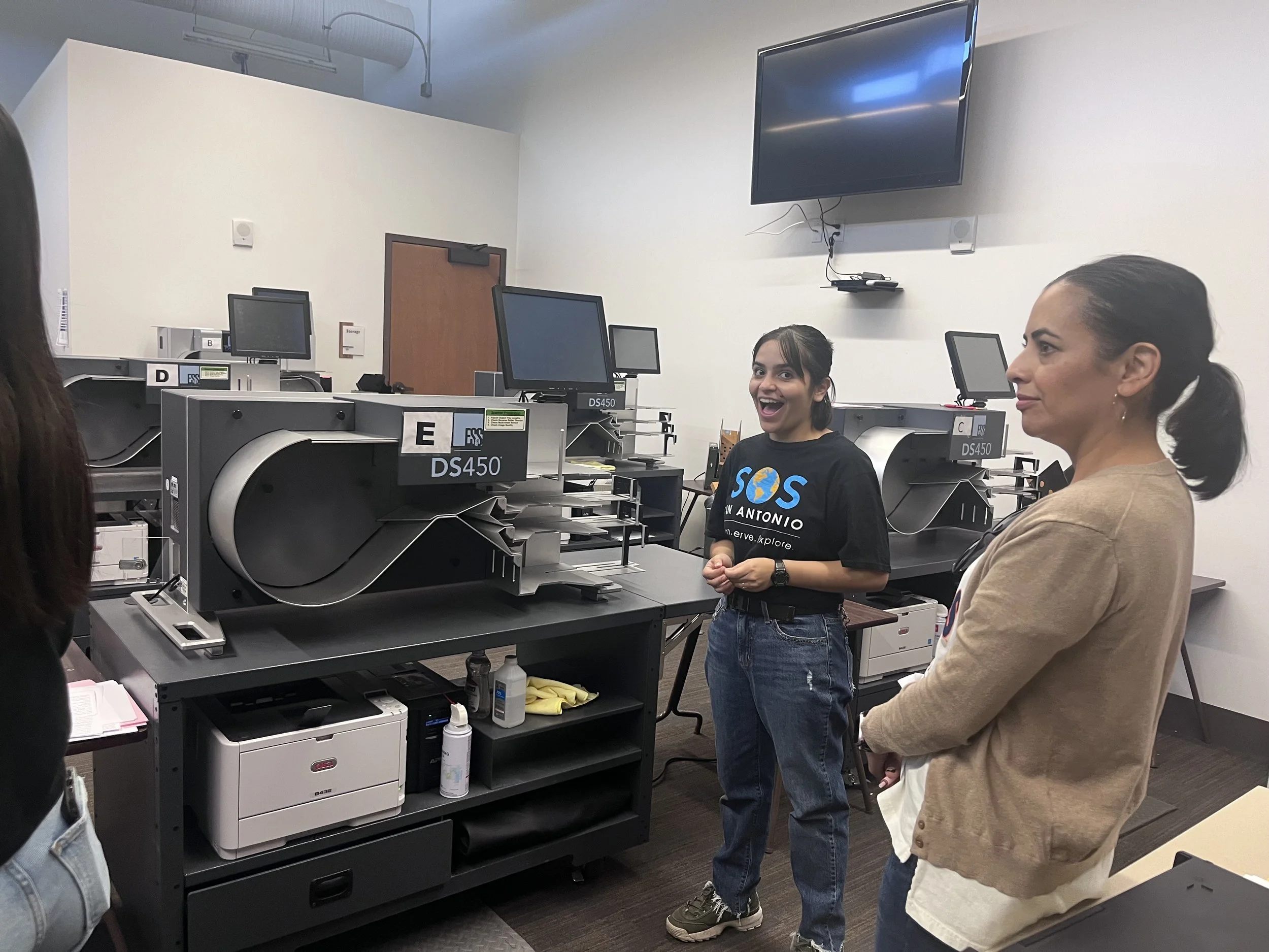 Two women standing in a classroom with computers and machines, one woman smiling and talking, the other listening with arms crossed.