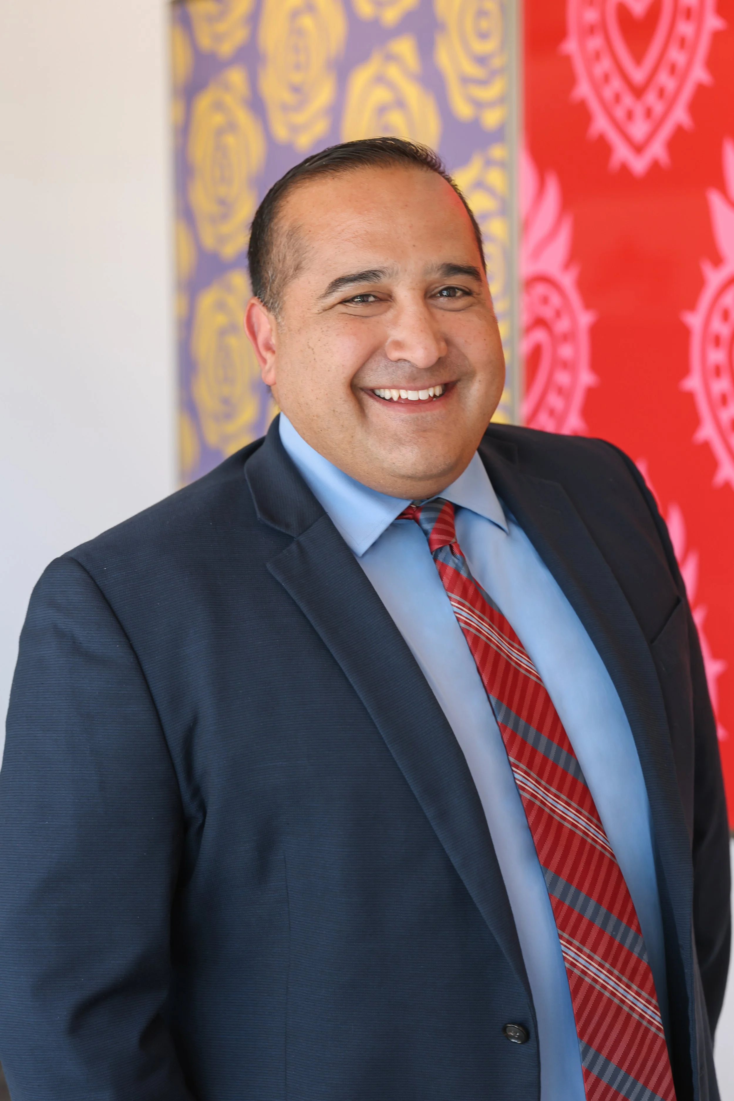 A smiling man in a navy suit, light blue shirt, and red plaid tie stands in front of colorful artwork, including purple with yellow roses and red with heart-shaped patterns.