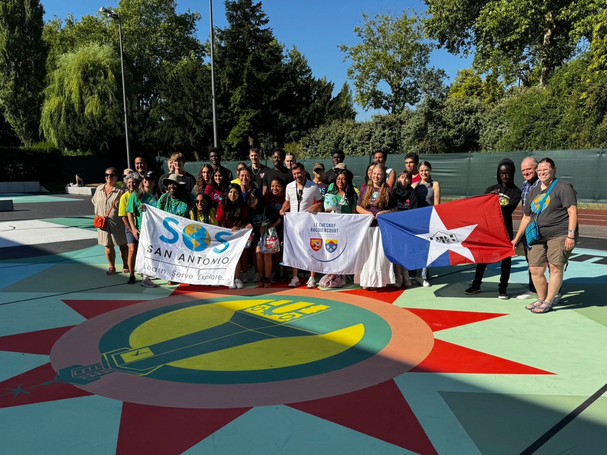 Group of people holding banners and flags on colorful outdoor basketball court with greenery in the background.