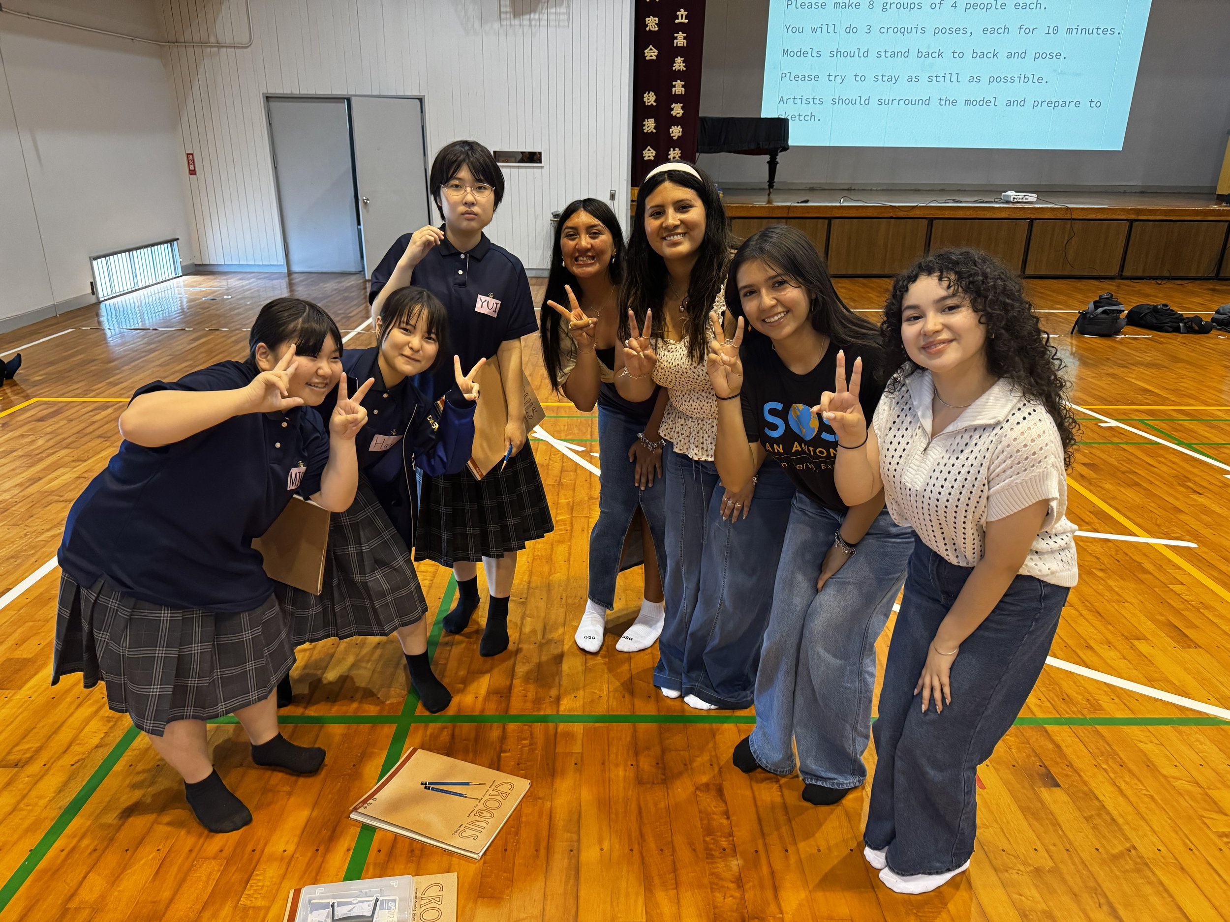 Group of seven diverse young students smiling and making peace signs in a gymnasium, with a stage and projection screen displaying text in the background.