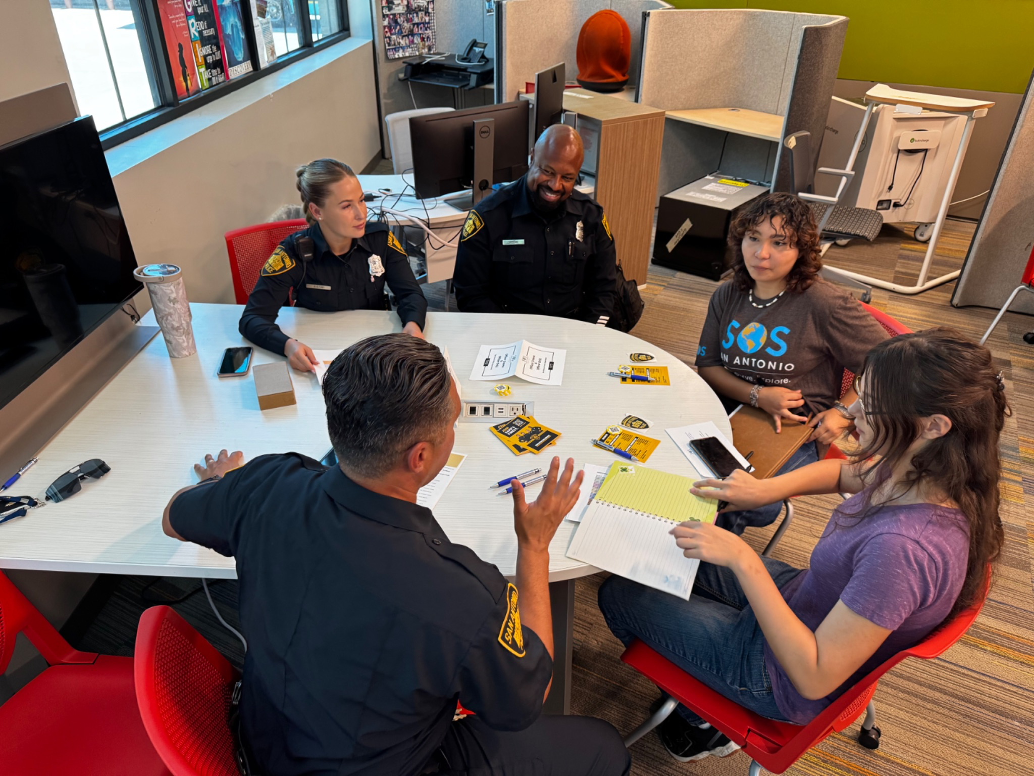 Group of five people, including firefighters and young women, seated around a white table in an office, engaging in discussion with pamphlets, notepads, and smartphones on the table.
