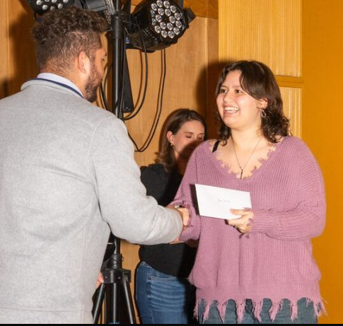 A woman in a pink sweater receiving a certificate and shaking hands with a man at an award or recognition event, with another woman smiling in the background.