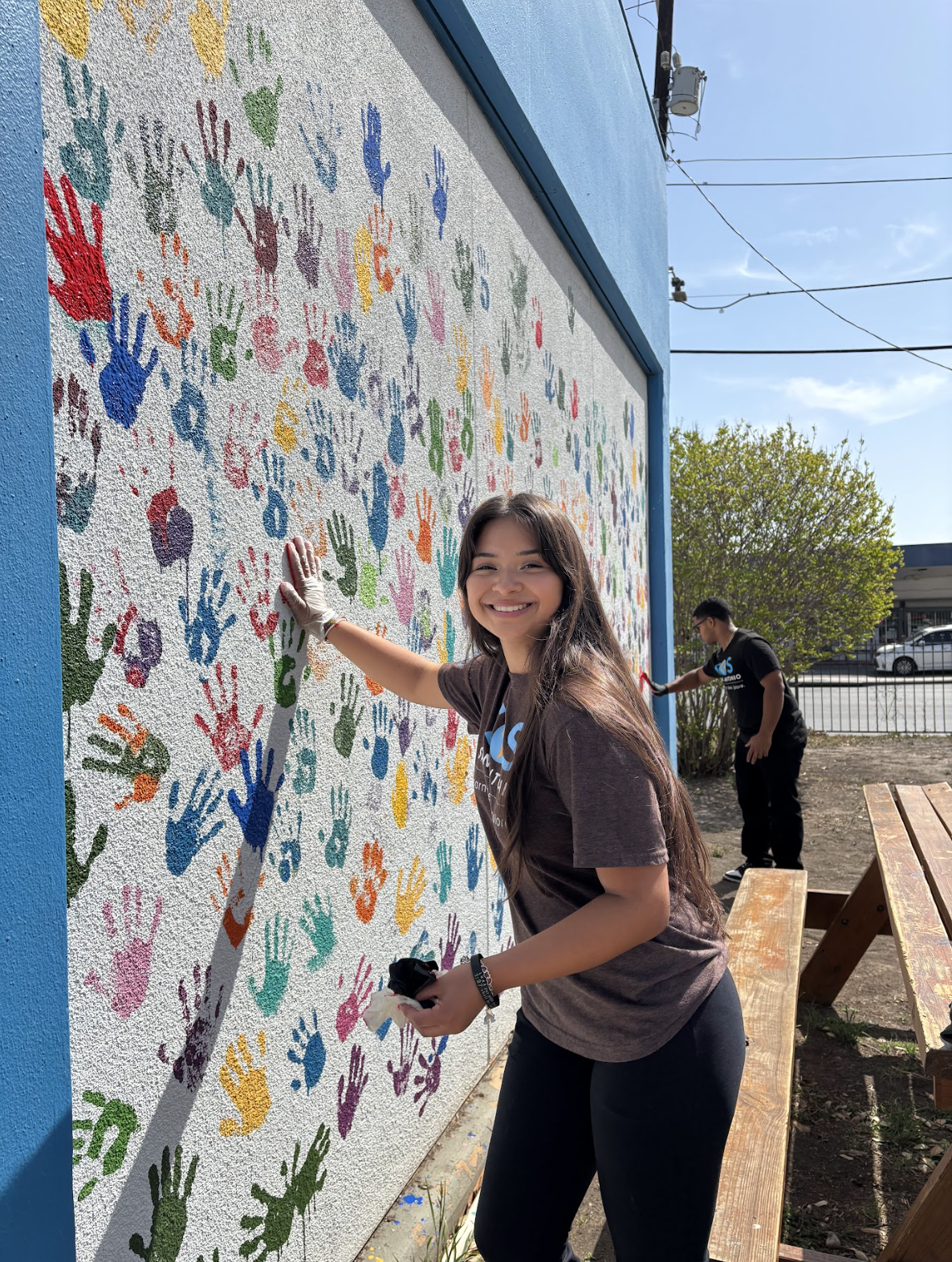 Young woman smiling while participating in a community mural painting event, creating colorful handprints on a white wall outside on a sunny day.