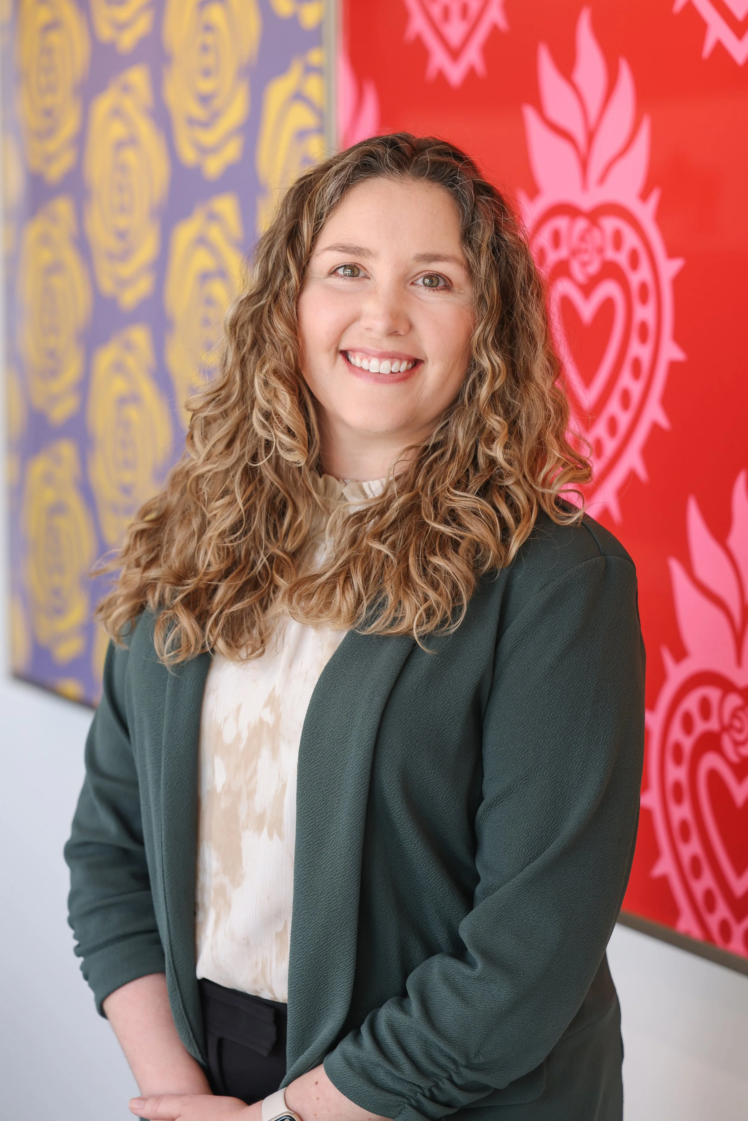 A woman with curly blonde hair smiling, standing in front of colorful artwork with heart and floral designs.