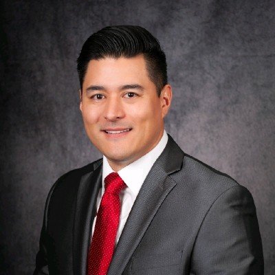 Professional headshot of a man in a gray suit and red tie, smiling, against a dark textured background.