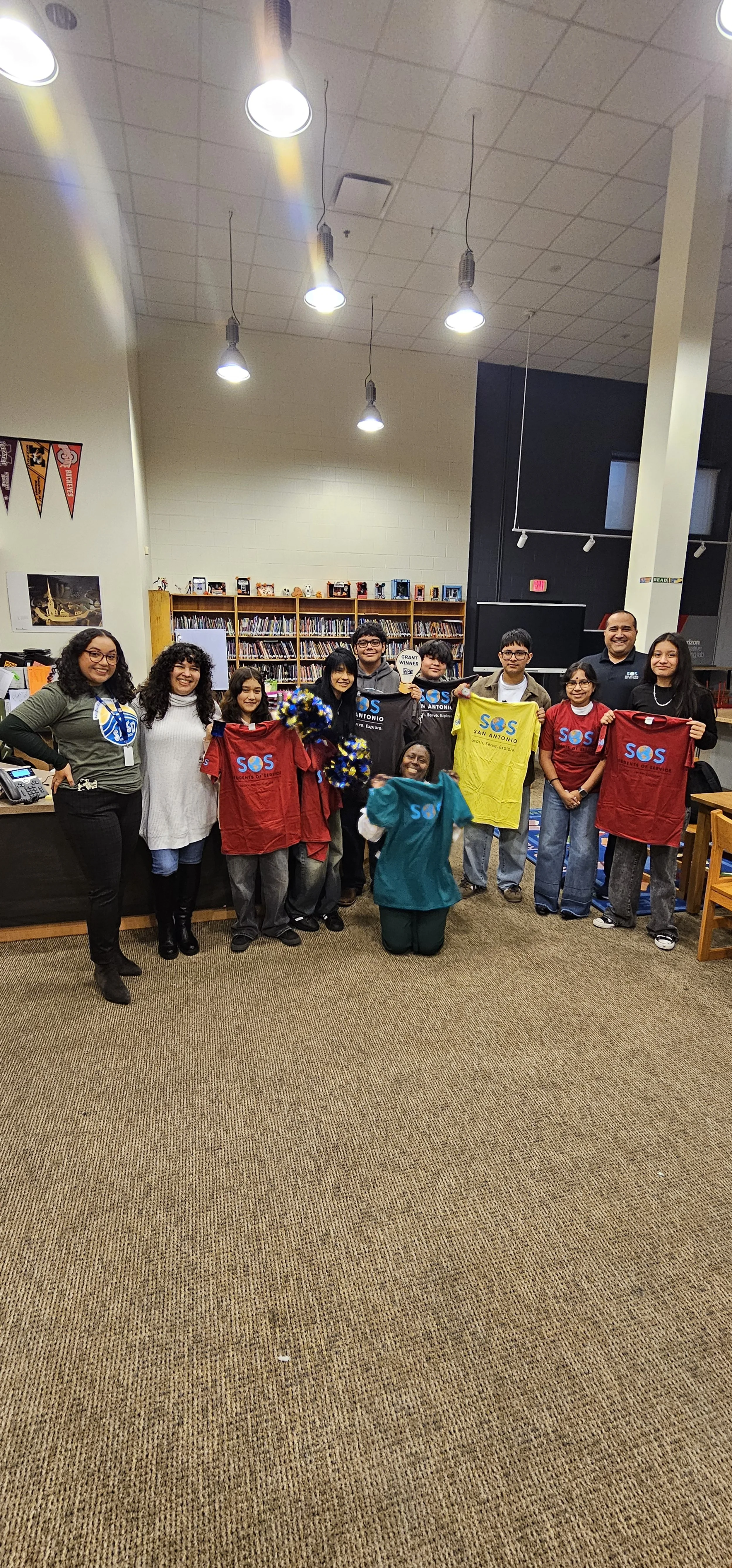 Group of people in a library holding SOS San Antonio shirts and cheerleading pom-poms.